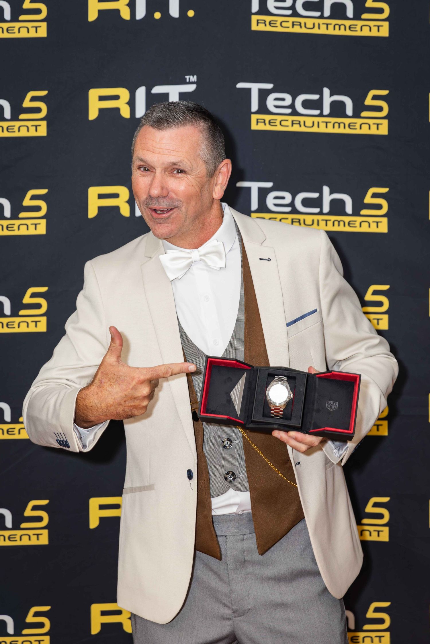 Man in cream blazer proudly displays a watch award at Tech 5 Recruitment event backdrop.