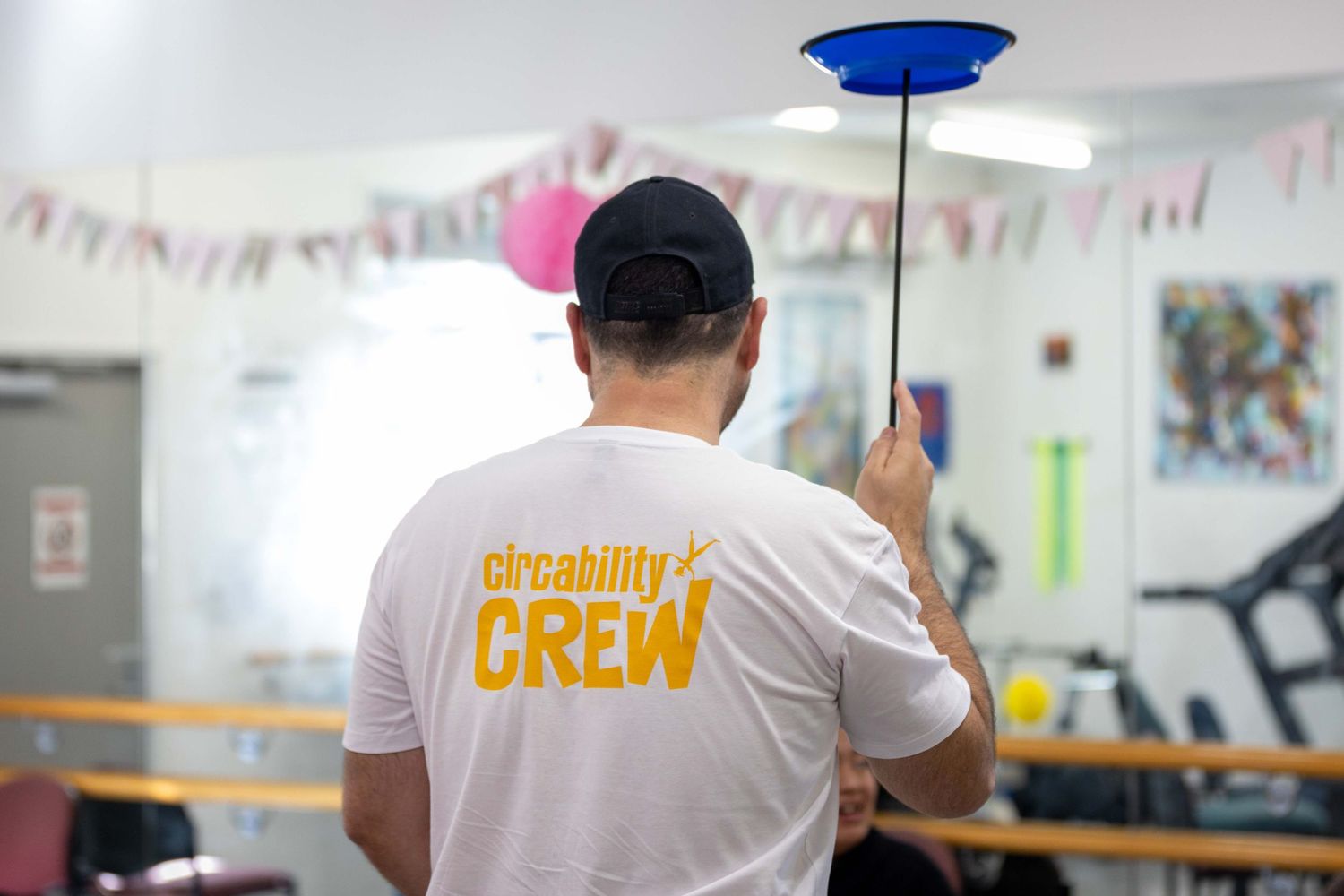 A person wearing a Circability Crew t-shirt spins a blue plate on a stick in a colorful gym.