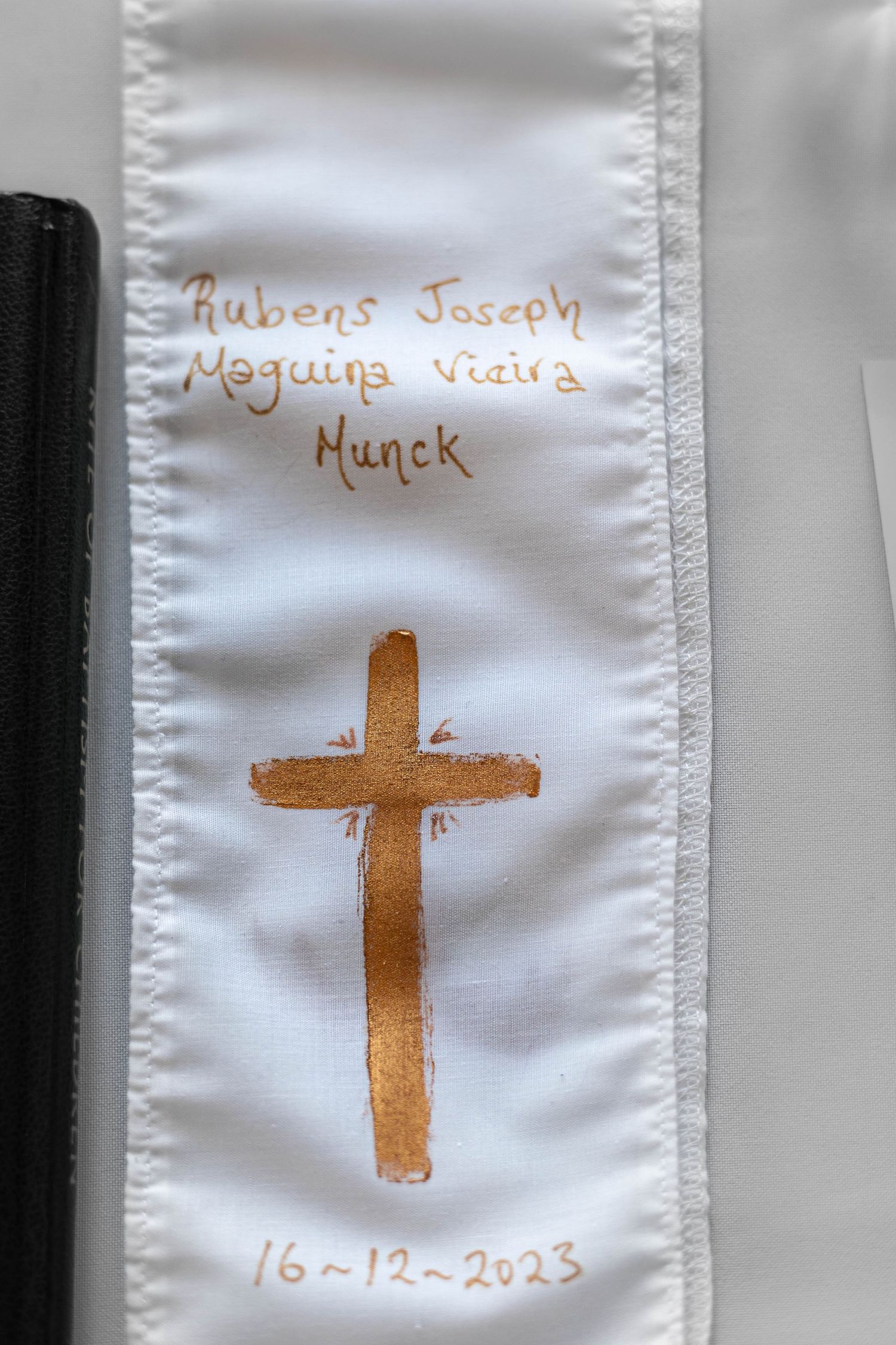 White fabric bookmark with gold cross and name Rubens Joseph Maguina Vieira Munck, dated 16-12-2023.