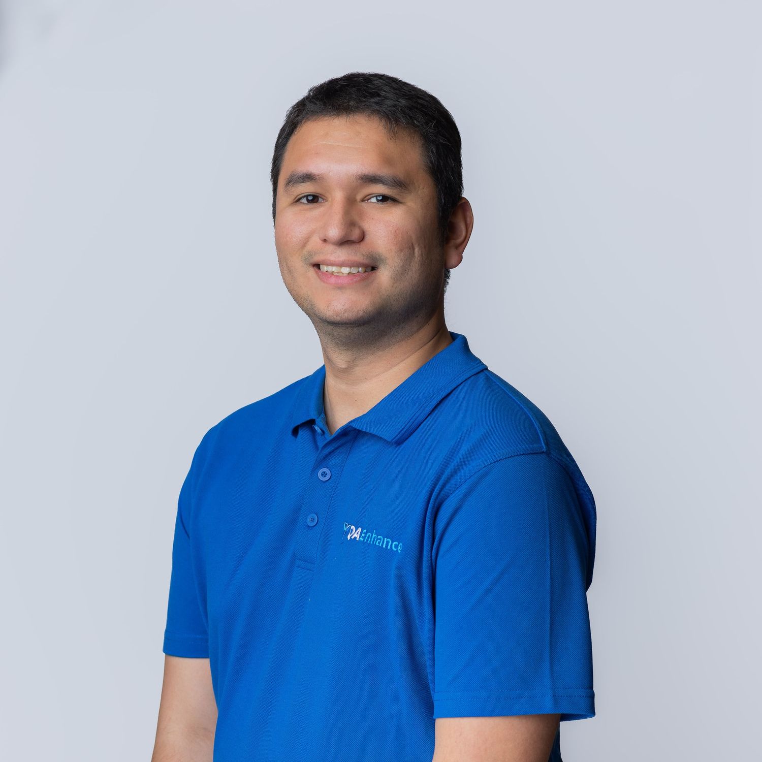 Young professional man smiling in a blue polo shirt against a light gray background.