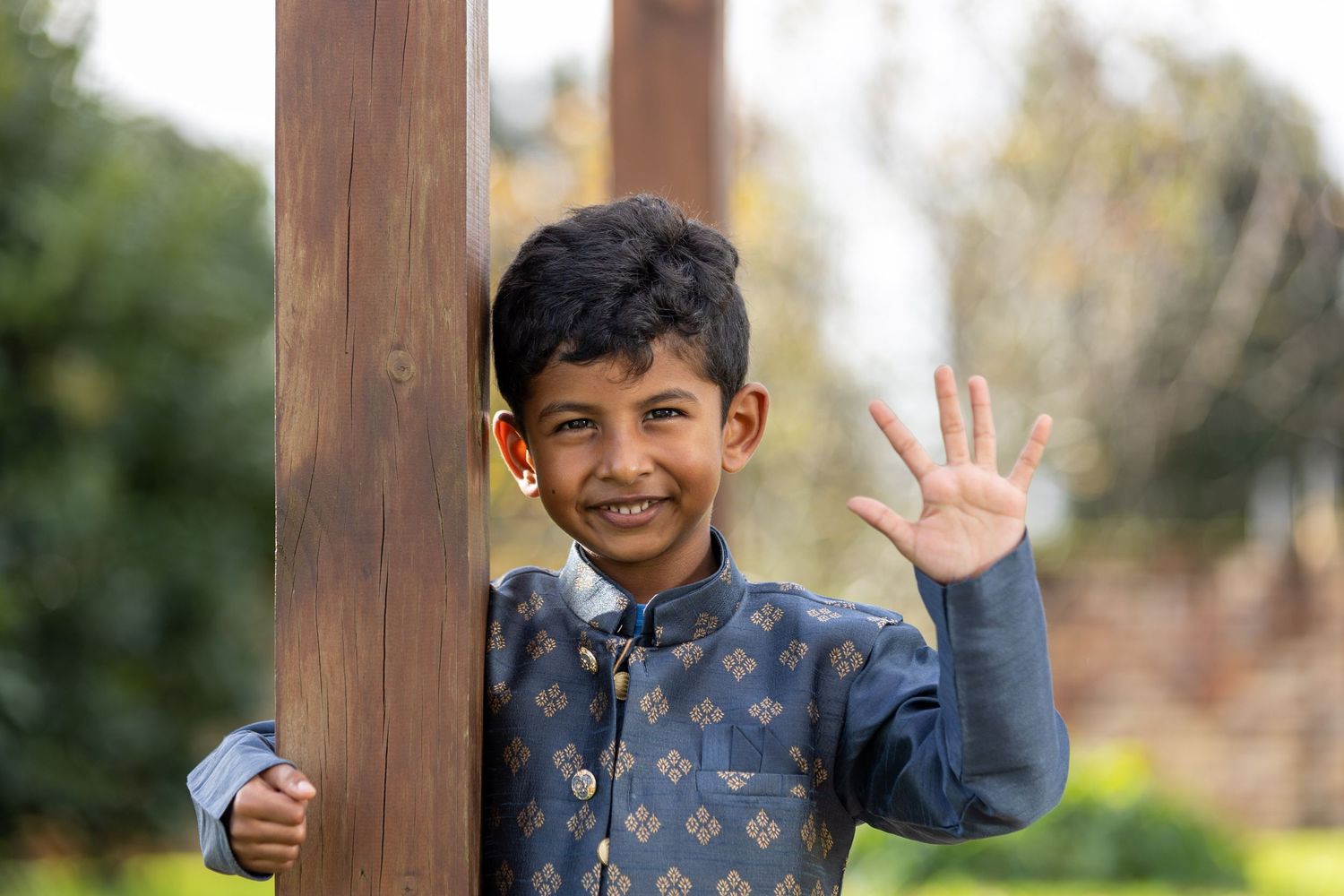 A smiling Indian boy in traditional blue kurta waves at the camera while holding a wooden post outdoors.