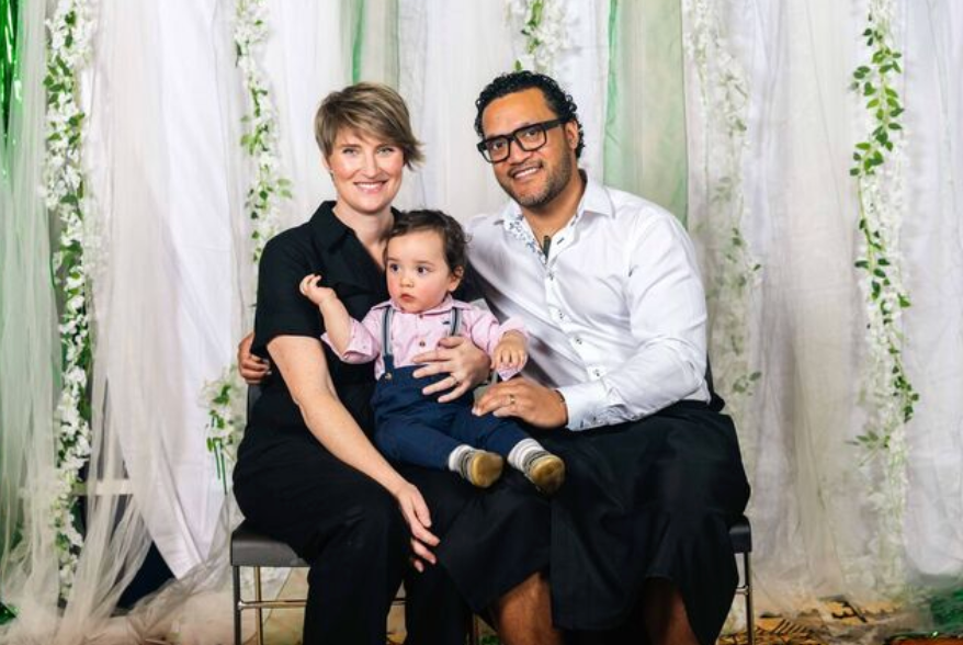 A smiling family of three poses for a portrait, with parents holding their baby dressed in a white shirt and suspenders.