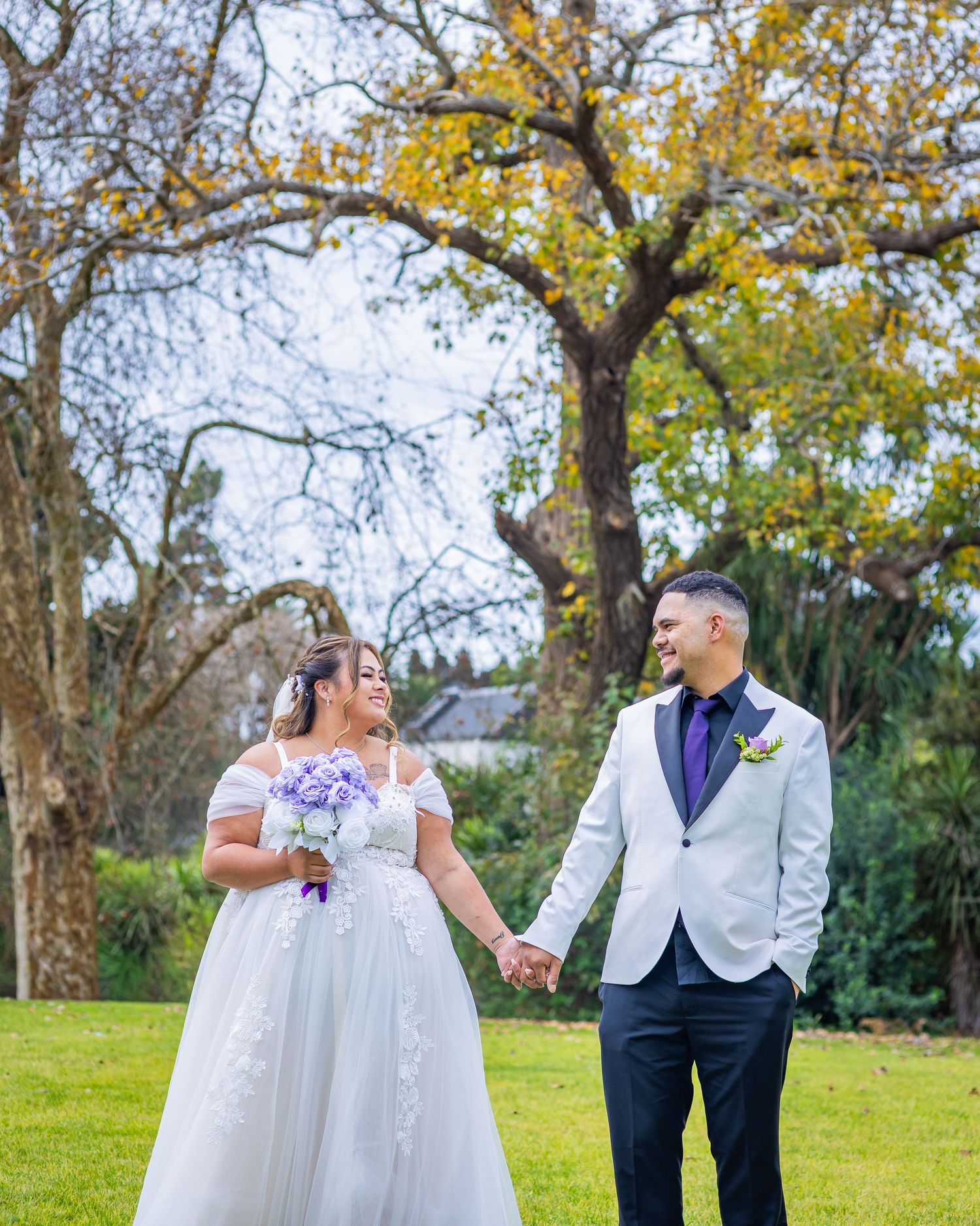 Bride and groom holding hands in a lush garden, she in a white floral gown, he in a white blazer with purple tie.
