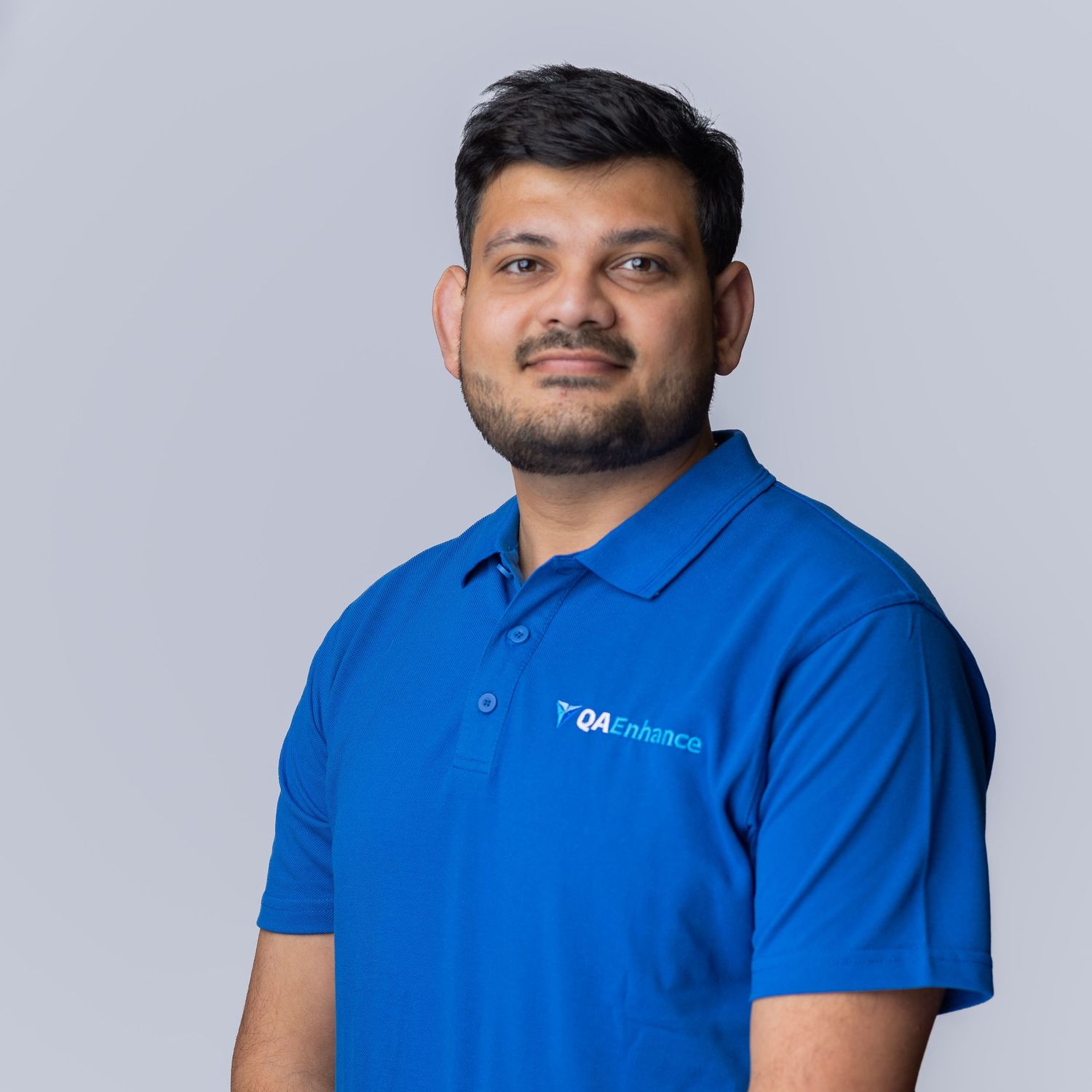 Professional man in blue QAEnhance polo shirt smiling against gray background.