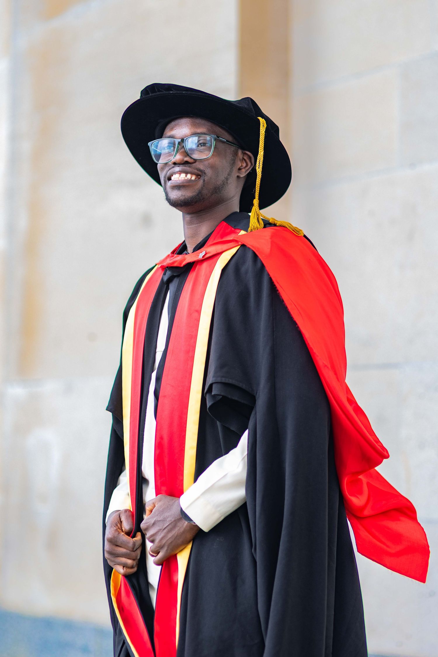 Smiling man in black doctoral graduation gown with red cape and mortarboard hat posing outdoors.