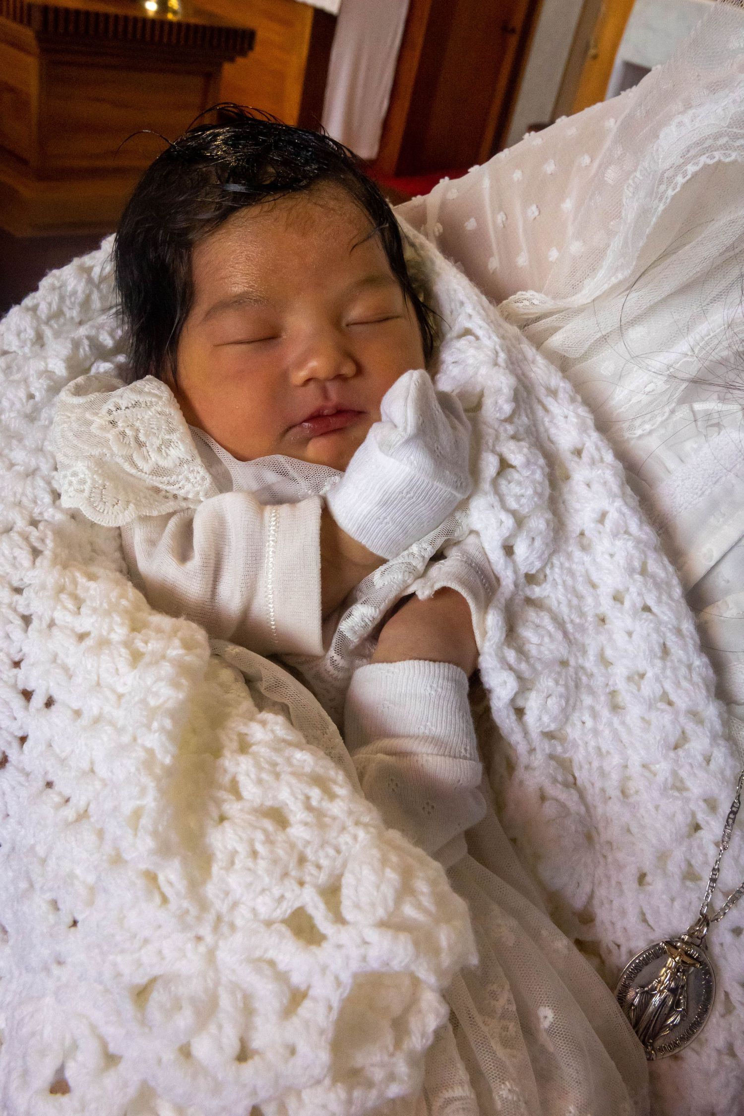 Newborn baby sleeping peacefully in white christening gown, lace blanket, and mittens with religious medallion.