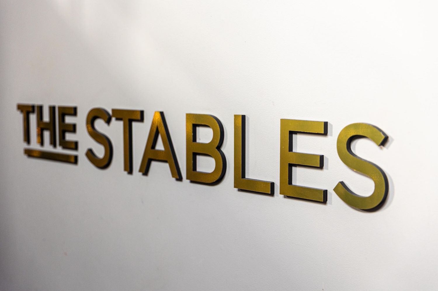 Gold metallic letters spelling The Stables mounted on a white wall, elegant signage for a venue or business.