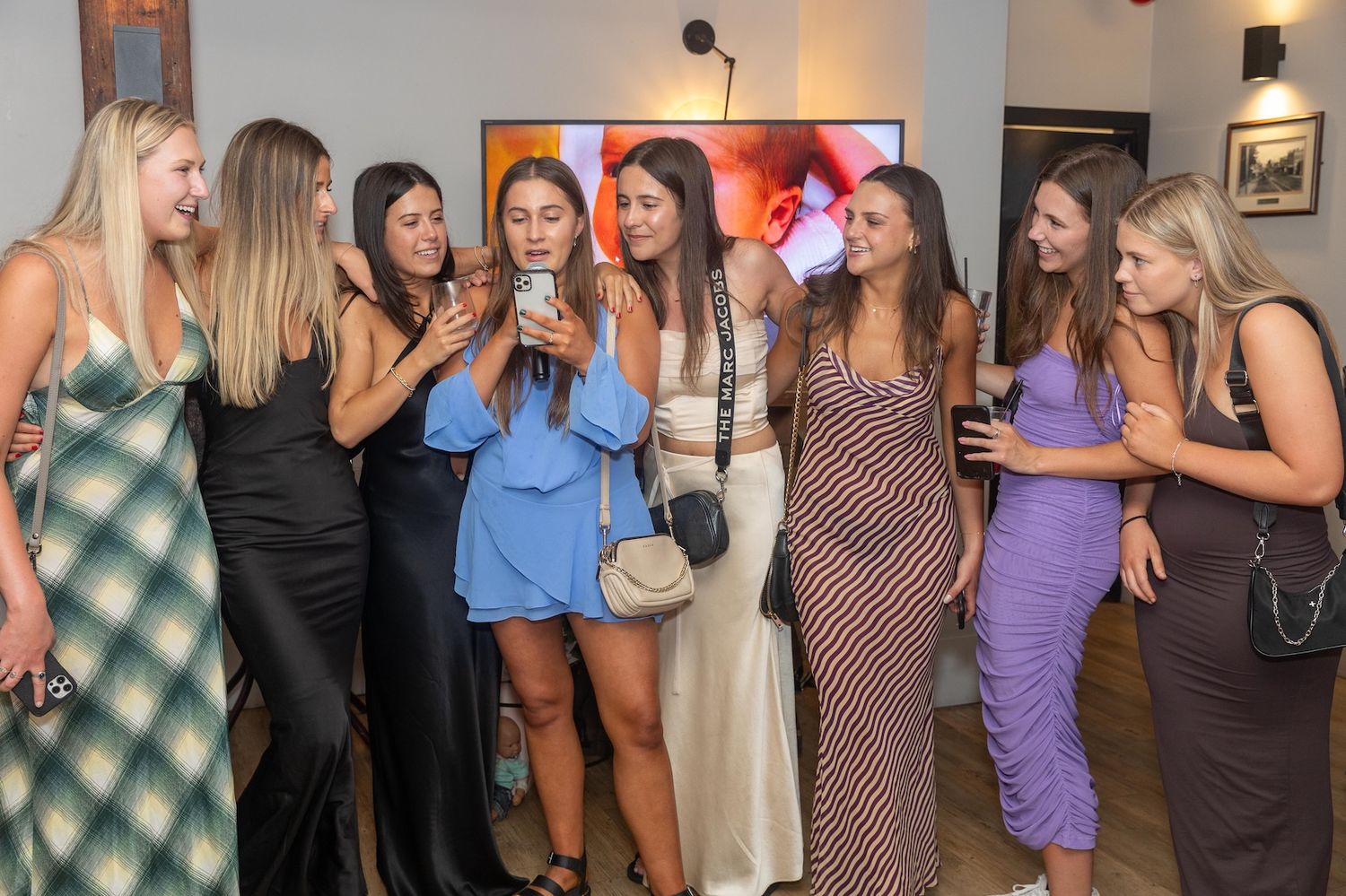 Group of eight women in elegant dresses gathered at a party, smiling and looking at a phone screen together.