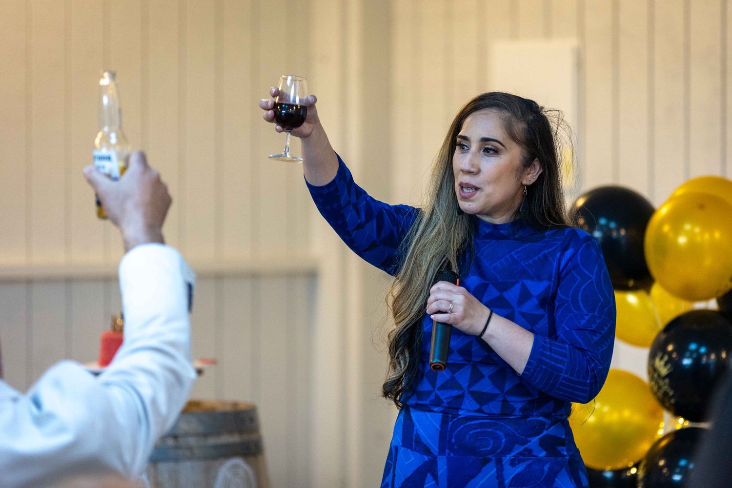 Woman in blue dress raising wine glass for toast at celebration with black and gold balloons.