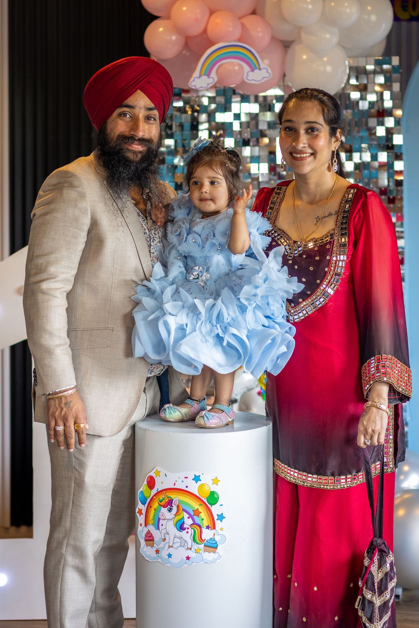 Sikh family posing at a rainbow unicorn birthday party, parents in traditional attire with toddler in blue dress.