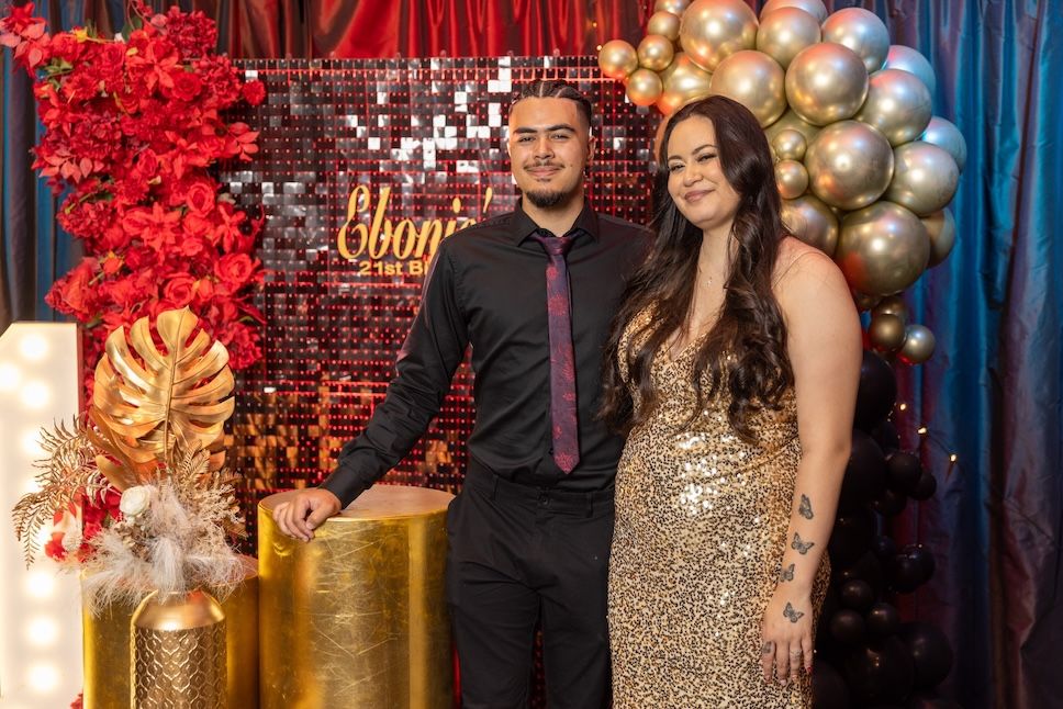 A couple poses together at a glamorous event with gold sequin backdrop, red floral decor, and balloon arch.