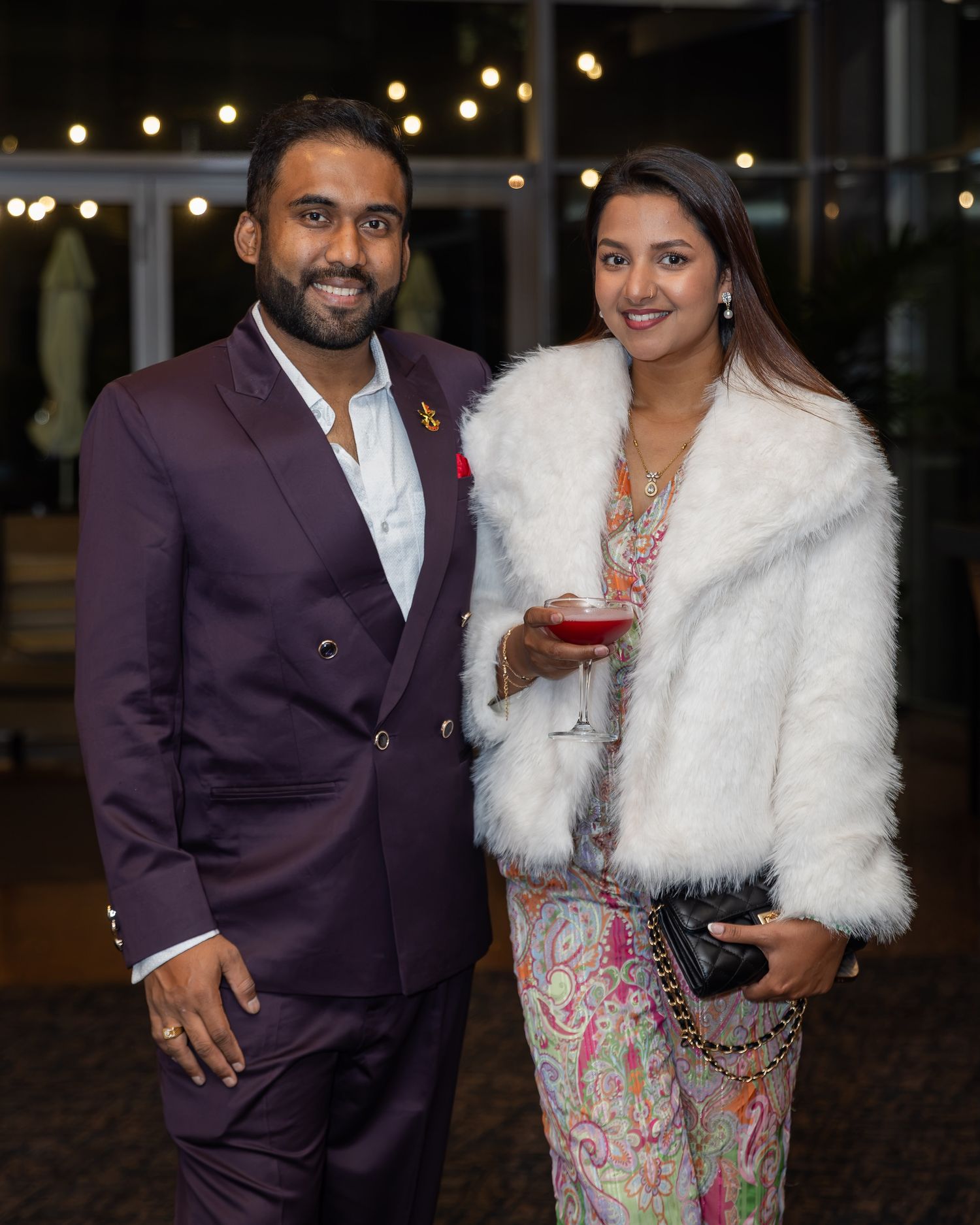 A man in a burgundy suit and woman in a floral dress with white fur jacket pose together at a formal evening event.