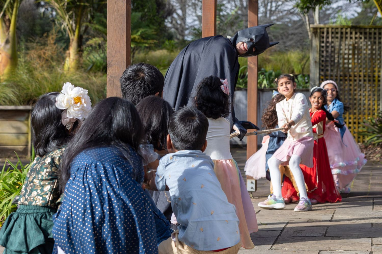 Children and adults in colorful traditional attire gather outdoors for a festive cultural celebration in a garden setting.