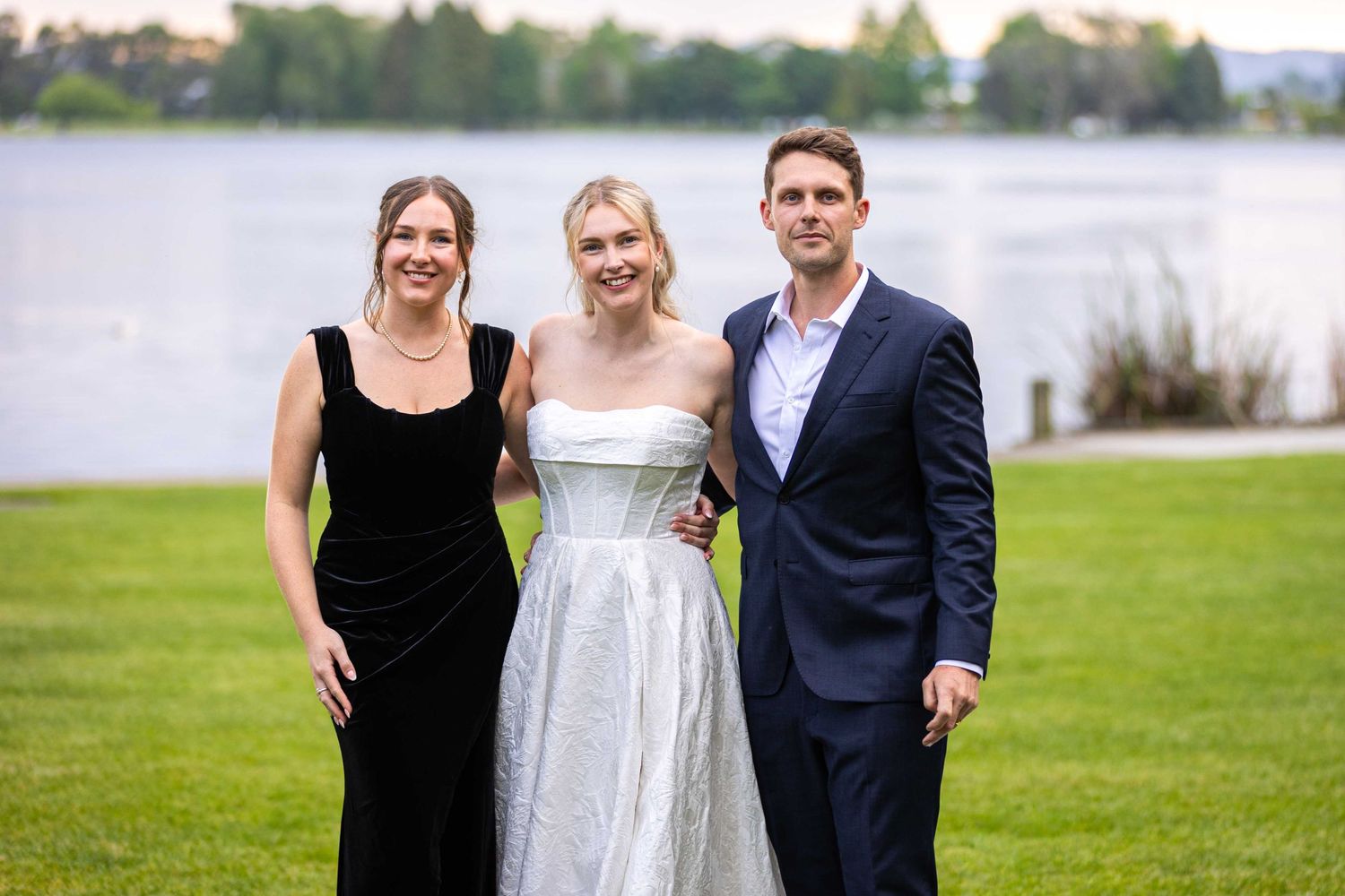 A wedding party poses together on grass with a lake backdrop.