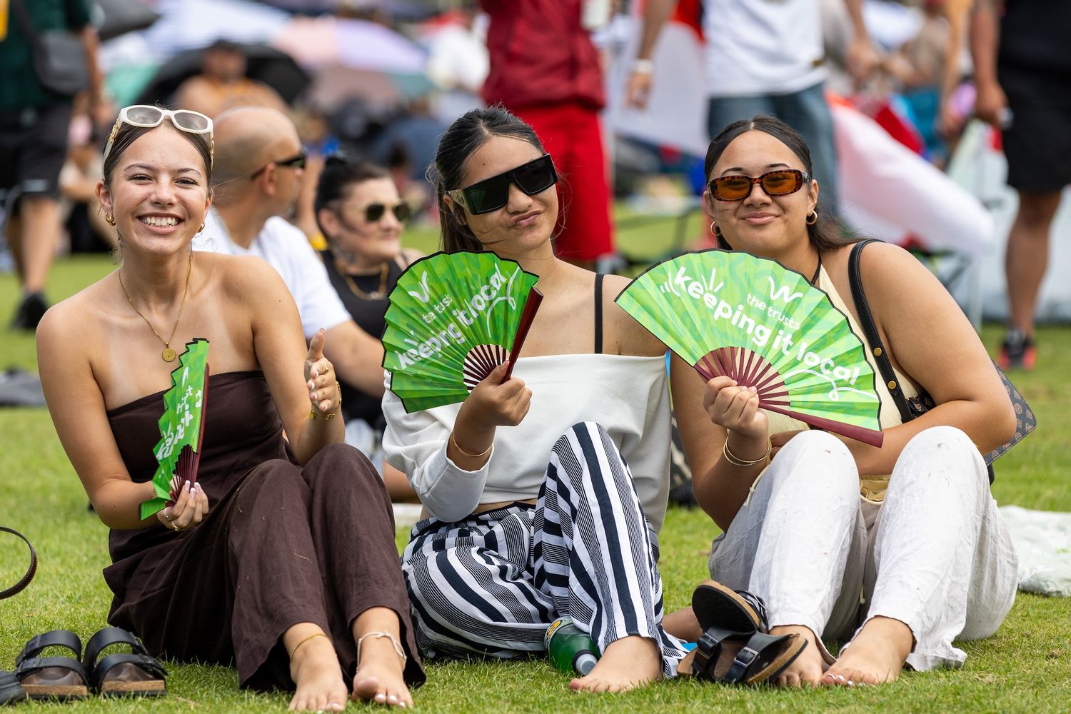 People lounging on grass holding green promotional fans at outdoor event.