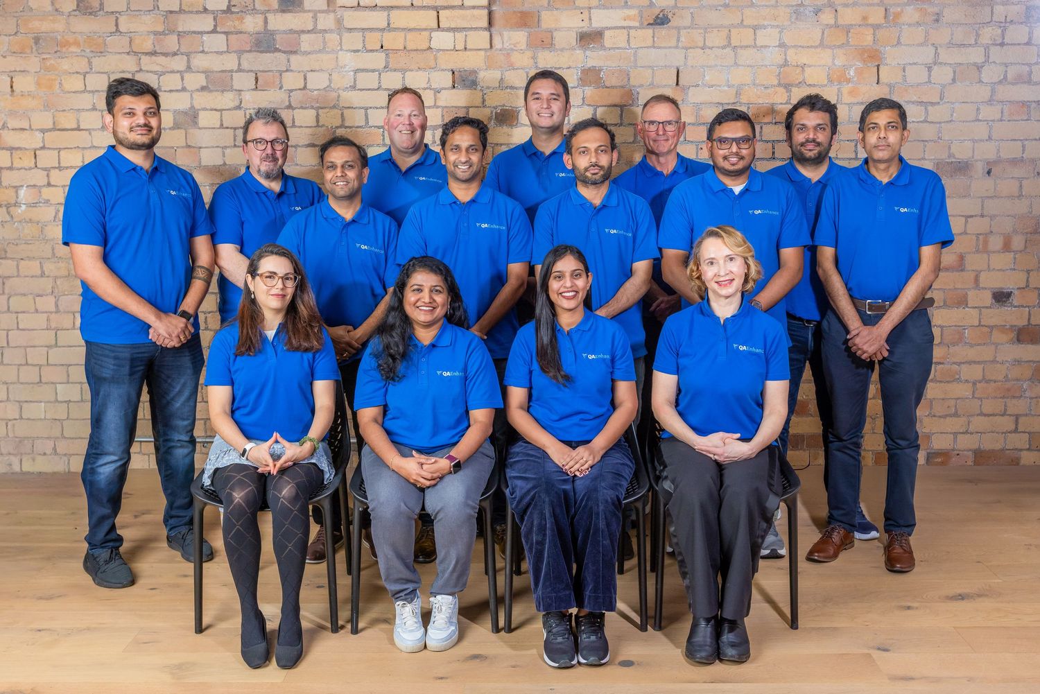 A professional team of diverse employees wearing matching blue polo shirts pose for a group photo in a modern brick-walled office.