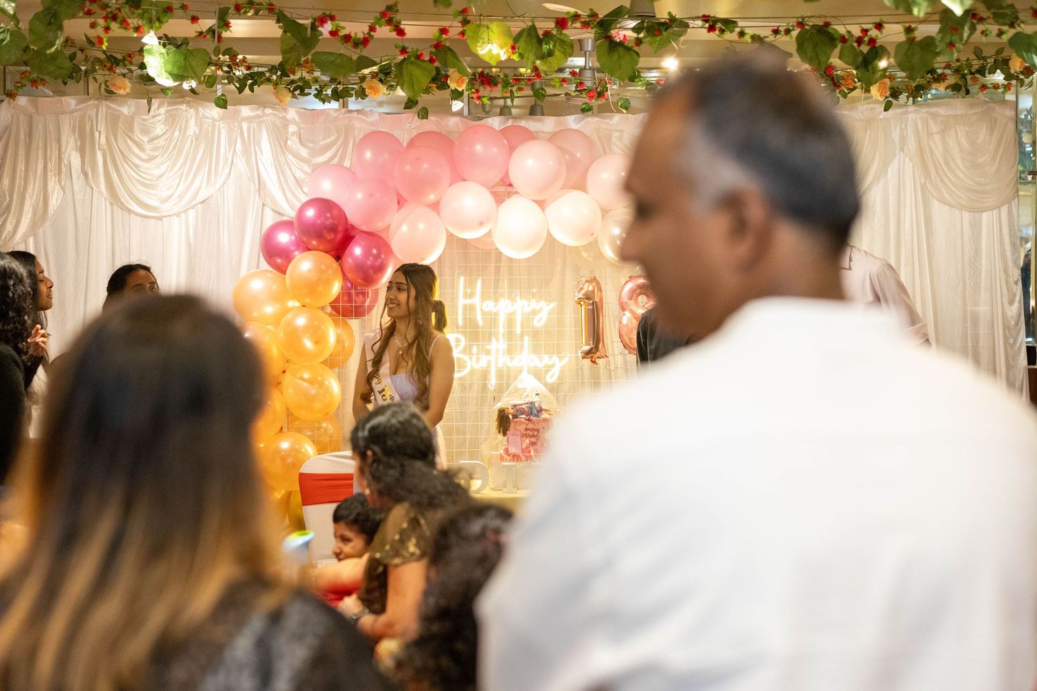 Colorful birthday party celebration with balloons, guests, and a Happy Birthday sign in a decorated venue.