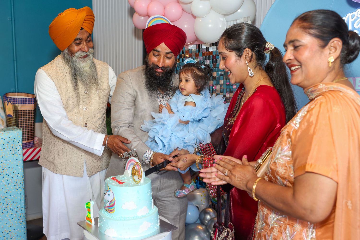 Sikh family celebrating a baby's first birthday with a decorated cake at a colorful party.