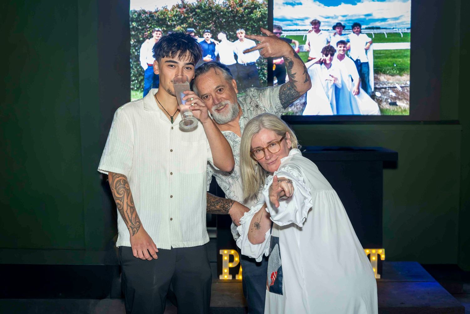 Three people smiling and posing for a selfie at an indoor event with a projected image in the background.