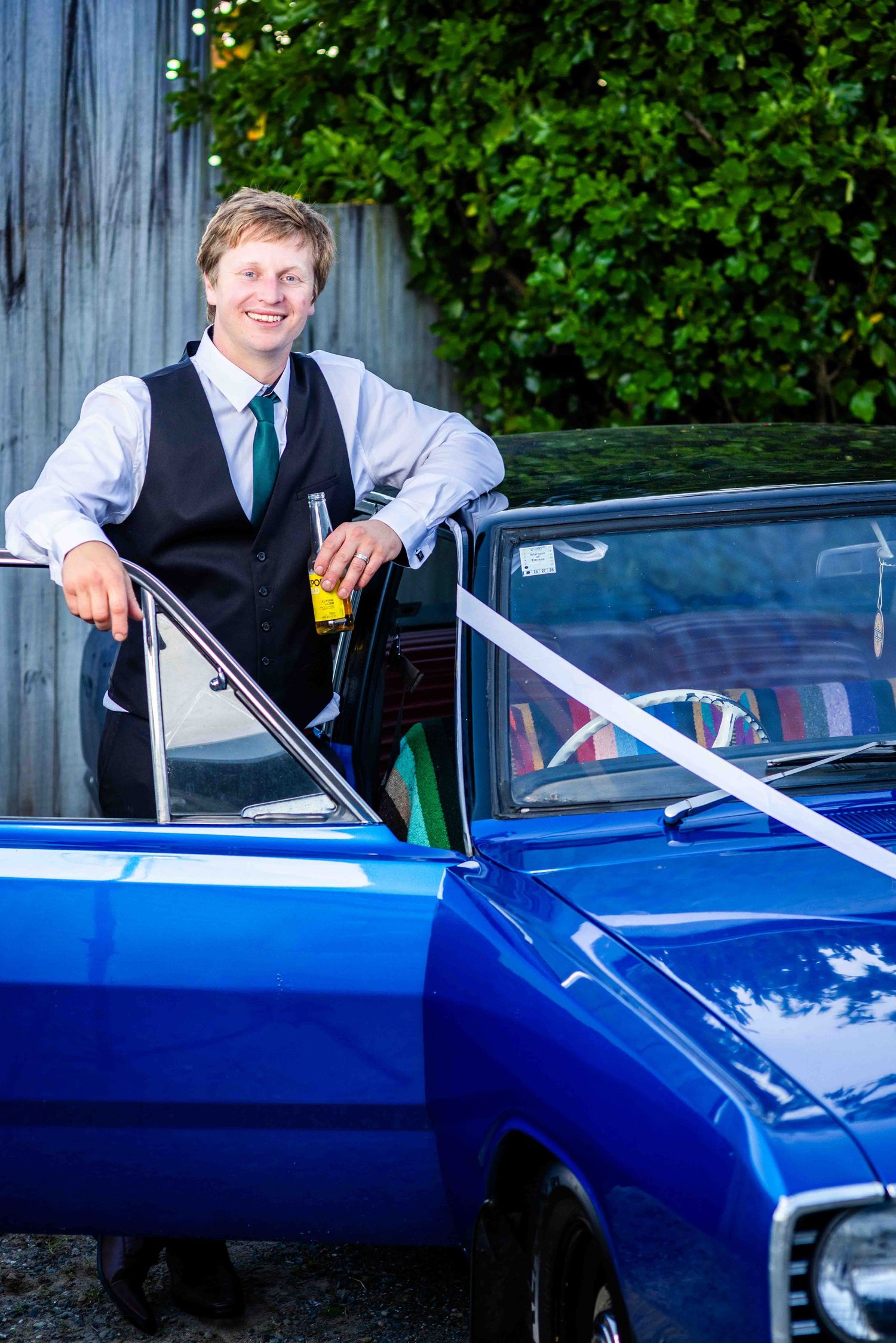 Smiling young man in vest and green tie leaning on a classic blue muscle car decorated with white ribbon outdoors.