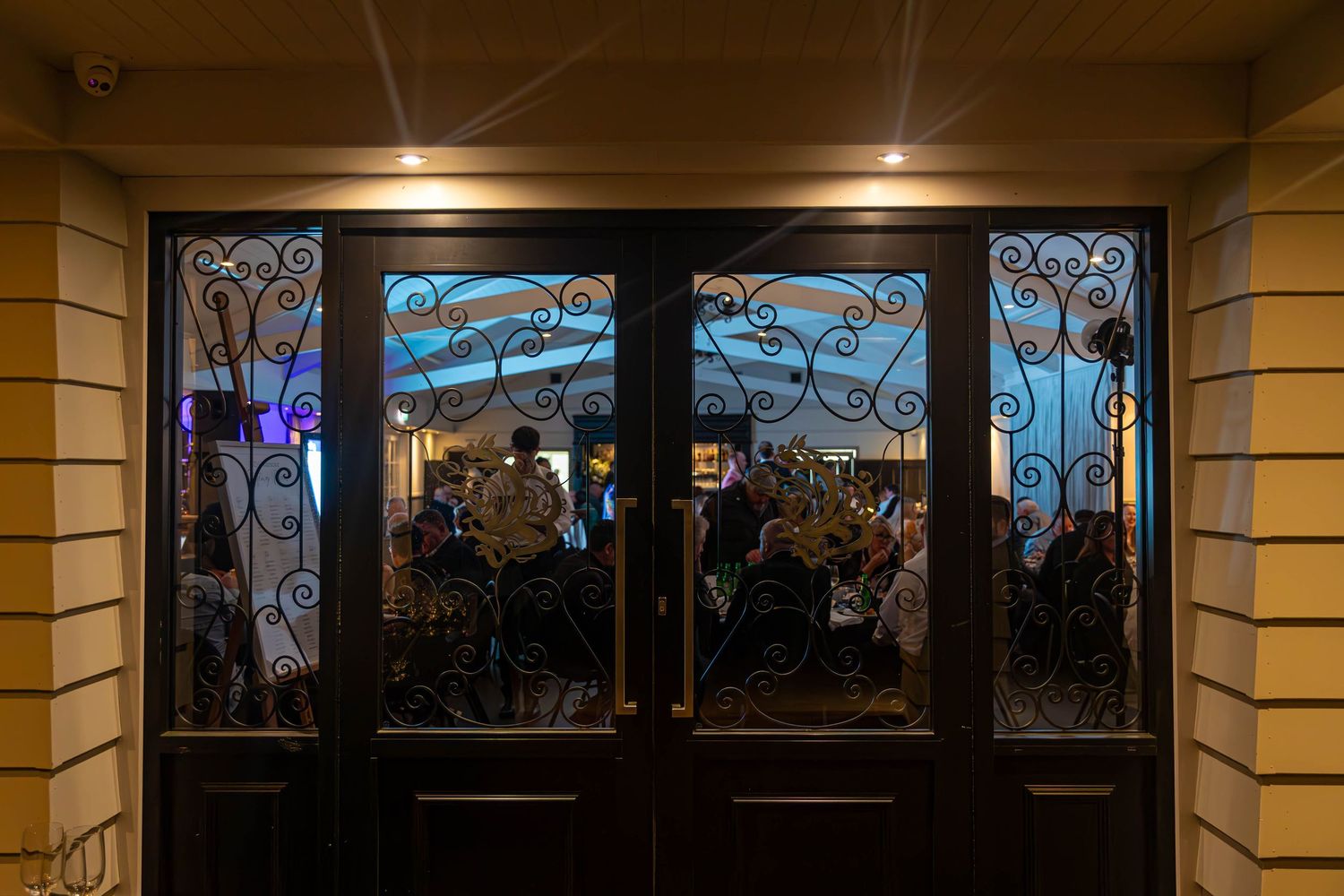 Ornate double doors with decorative wrought iron scrollwork reveal a warmly lit restaurant interior filled with diners.