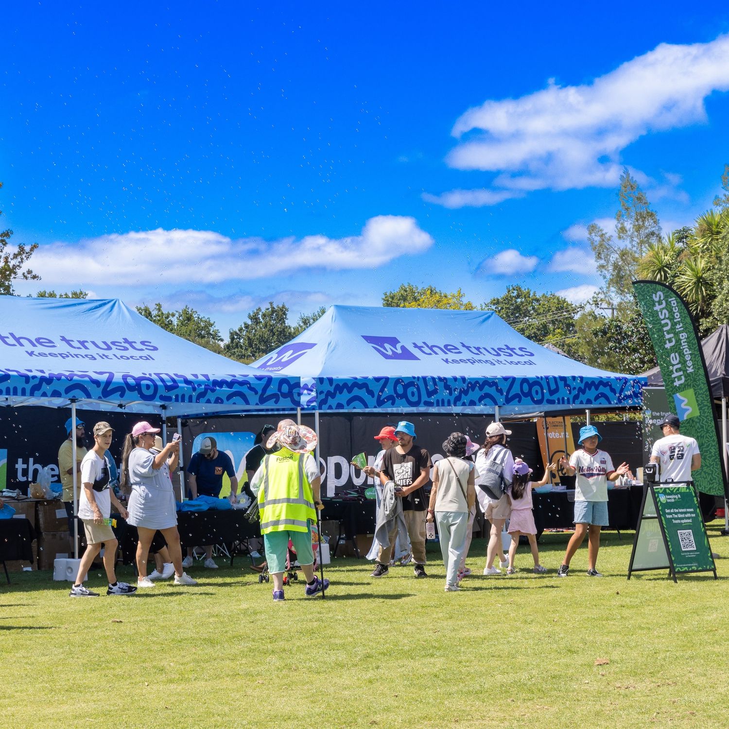 Crowd gathered under blue and white branded tents at outdoor summer event.