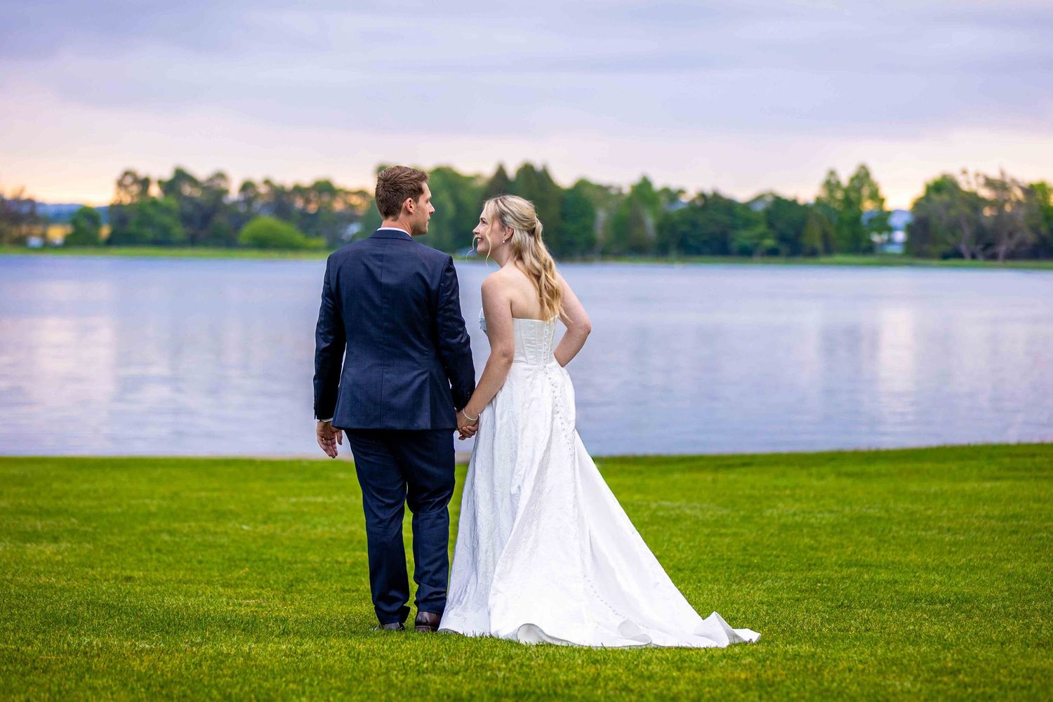 Couple walking along a scenic lakeside path at sunset in formal wedding attire.