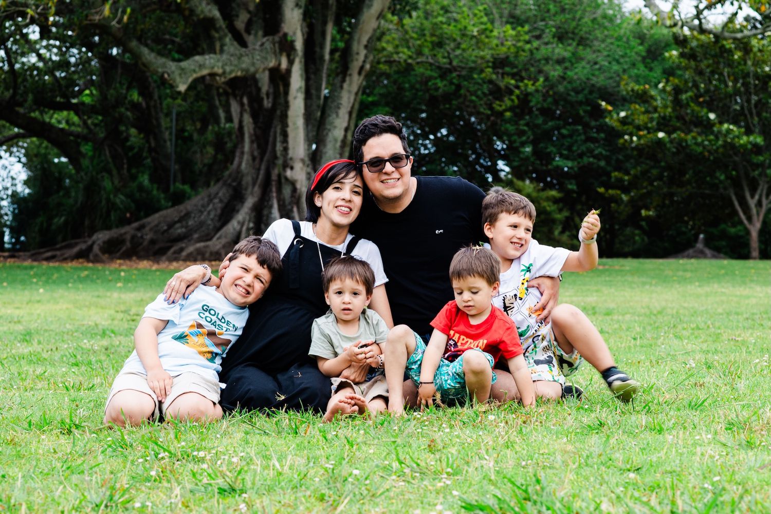Family of two adults and four children smiling together on green grass in a sunny park with large trees.
