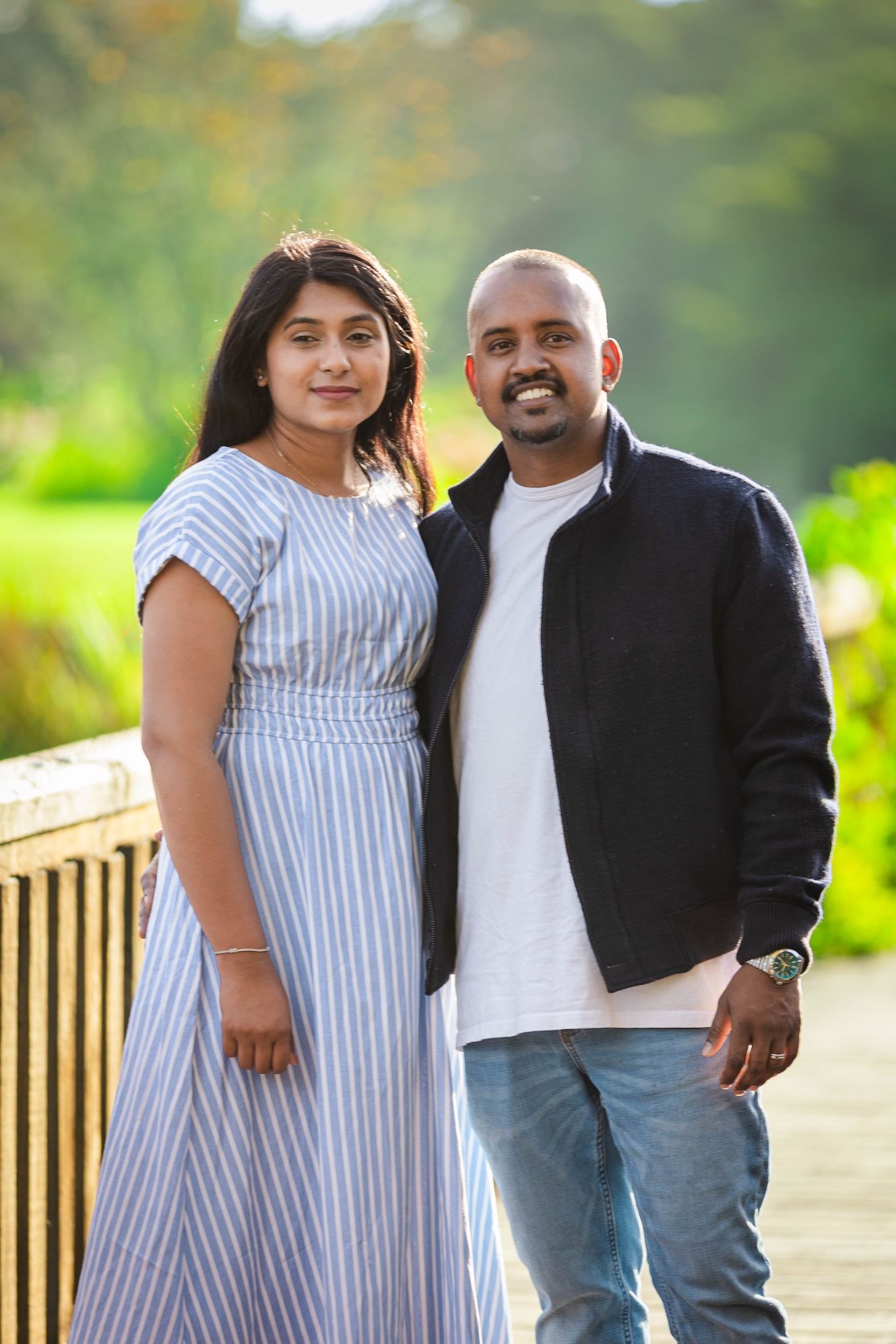 A smiling South Asian couple poses together on a wooden bridge surrounded by lush green trees.