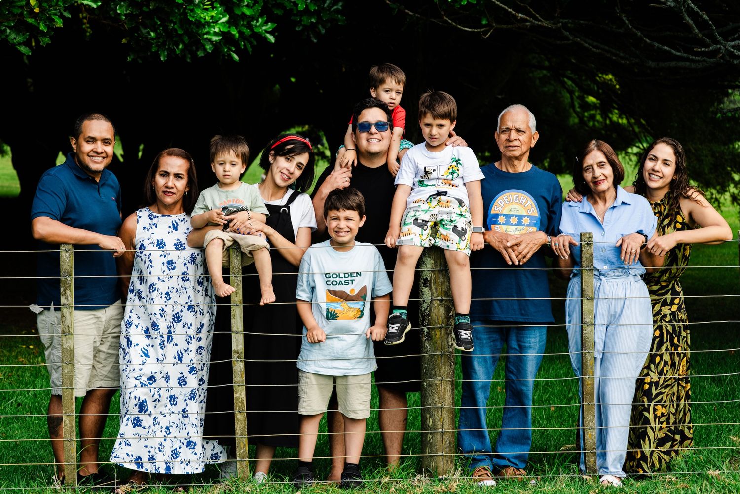 Large multigenerational family posing together outdoors by a fence surrounded by lush green trees.
