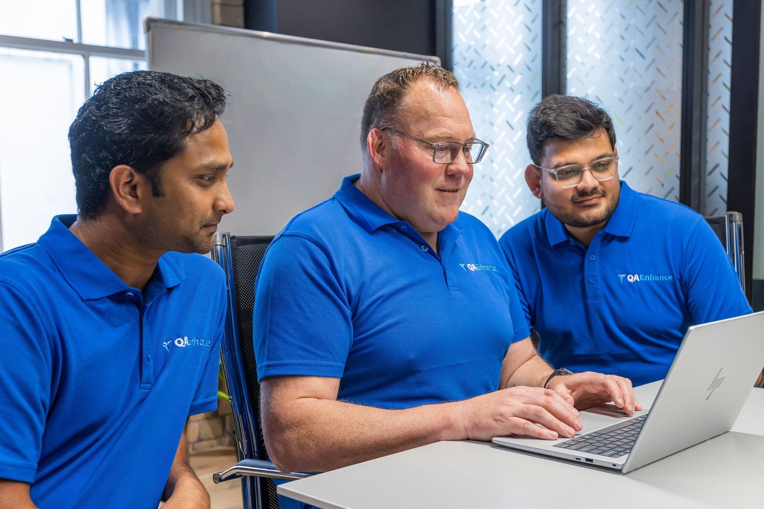 Three professionals in blue polo shirts collaborate on a laptop during a business meeting in a modern office.