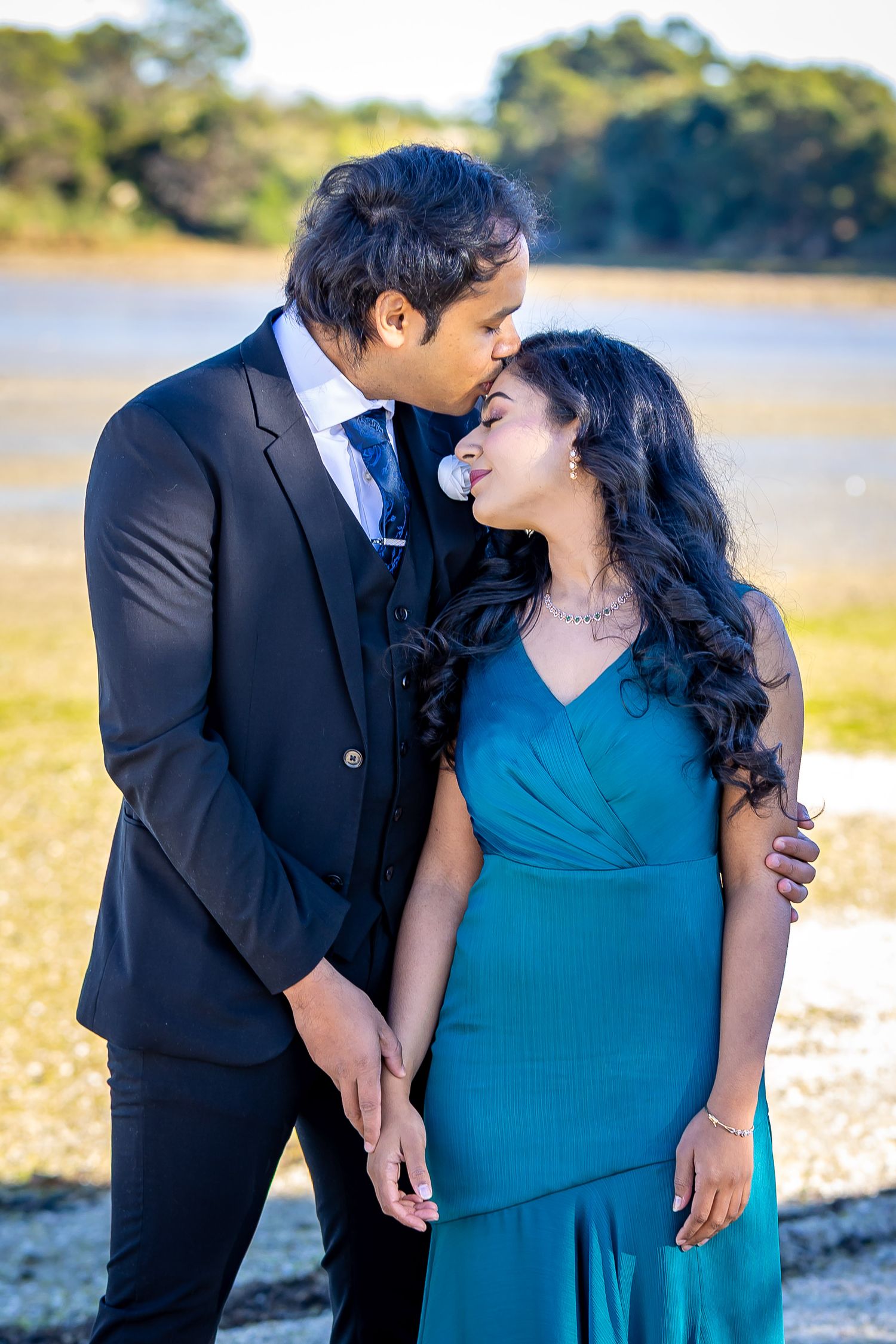 A couple in formal attire shares a romantic moment outdoors, man in black suit kisses woman in teal gown.