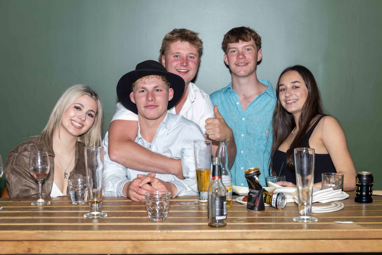 Five young friends smiling and posing together at a restaurant table with drinks and glasses.