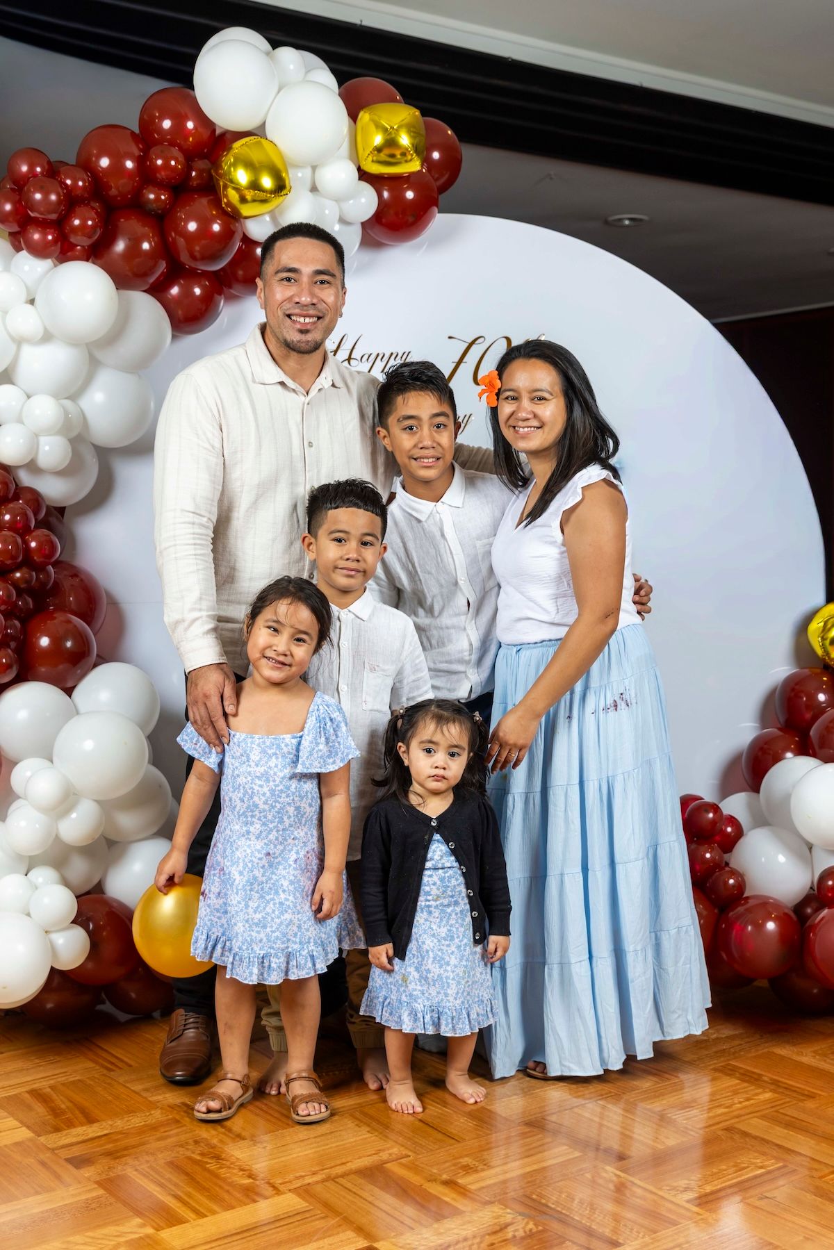 Family group wearing coordinated blue and white outfits poses in front of festive balloon arch decorations.