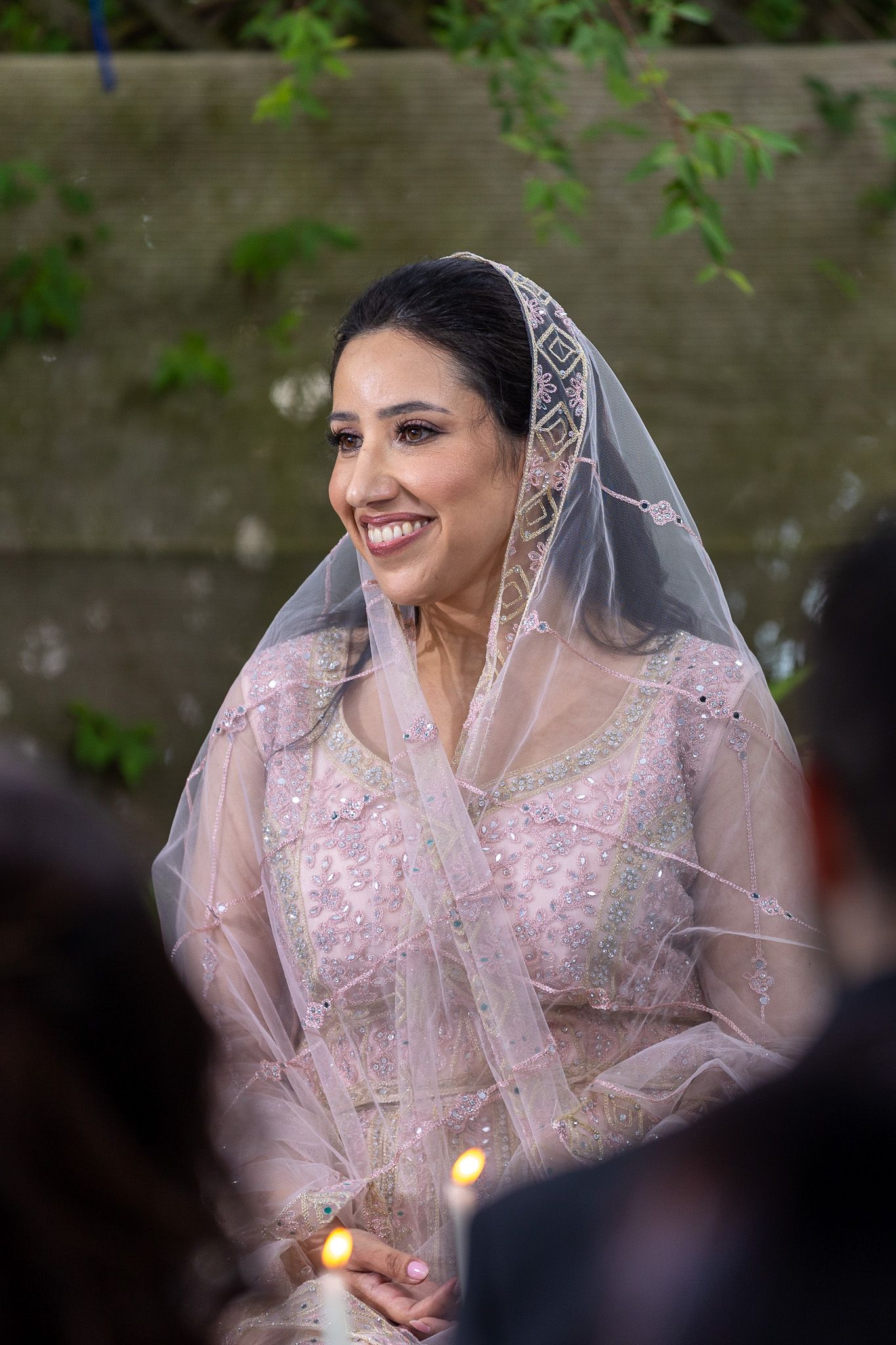 Smiling bride in pink embroidered lehenga with sheer veil holds candle at outdoor wedding ceremony.