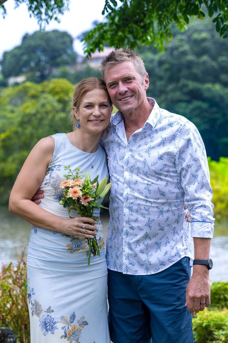 A smiling middle-aged couple poses outdoors near a lake, the woman in a floral dress holding a bouquet of peach flowers.
