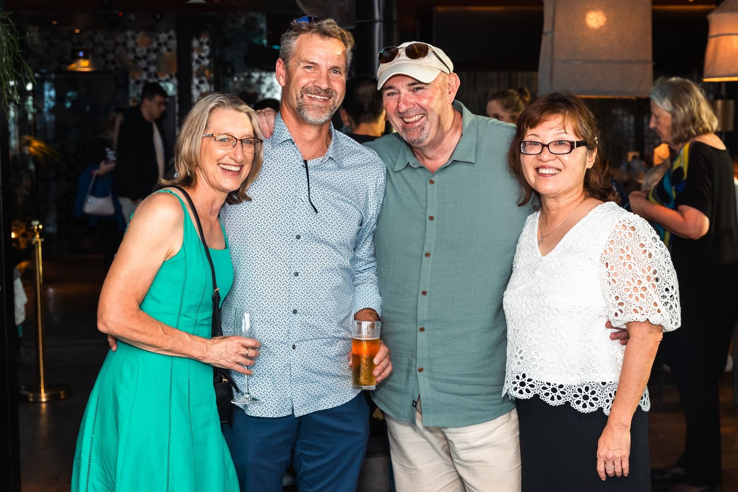 Four friends smiling and posing together at a social event, dressed in casual attire at an indoor venue.