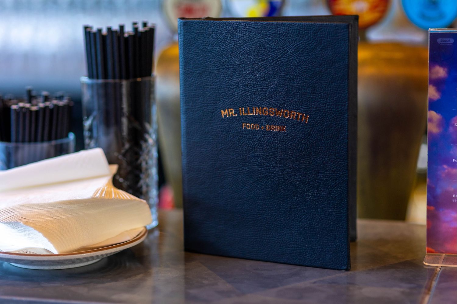 A navy blue book with gold lettering sits on a wooden table next to pens and plates.