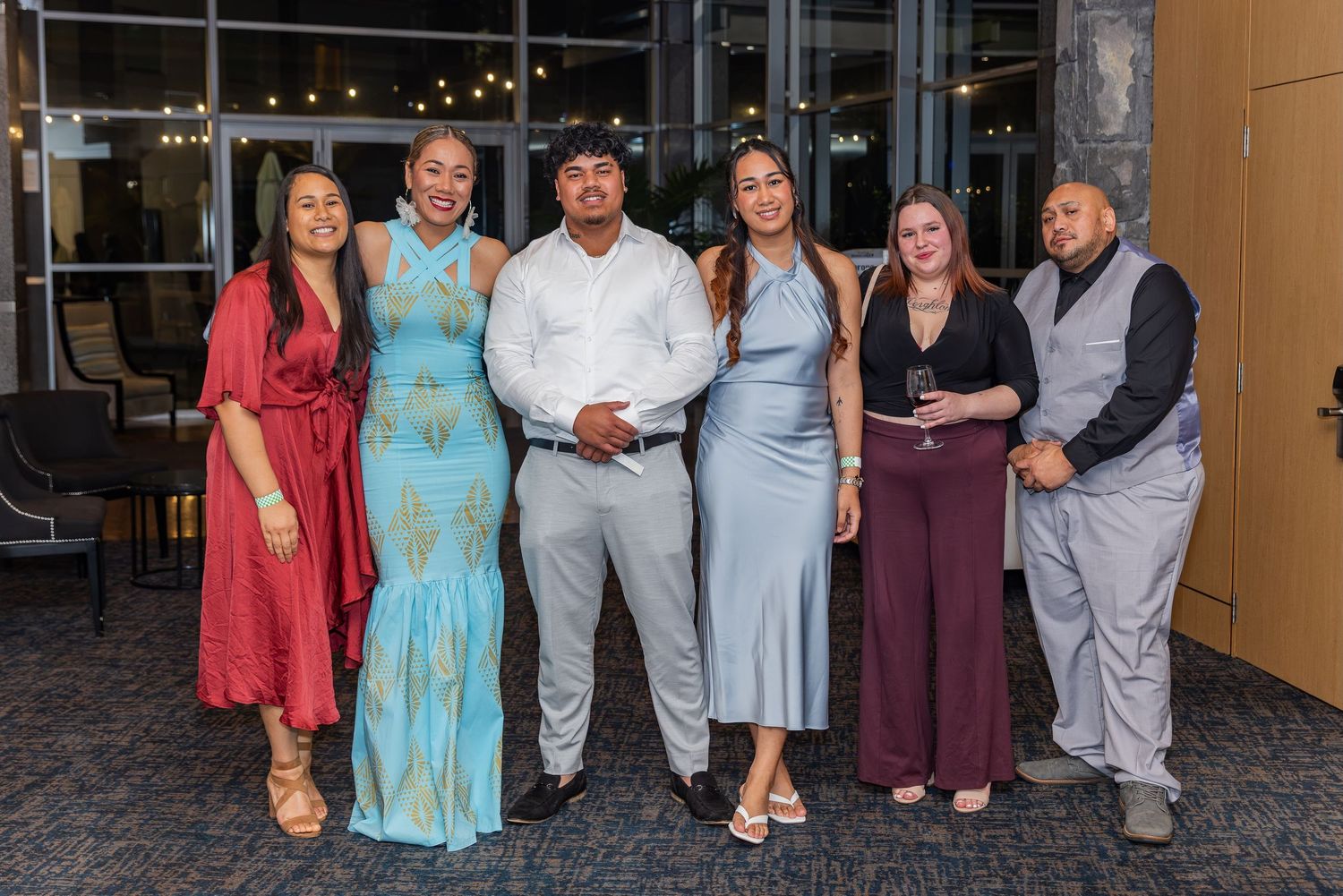 Group of six adults dressed in formal attire posing together at an elegant indoor event venue.