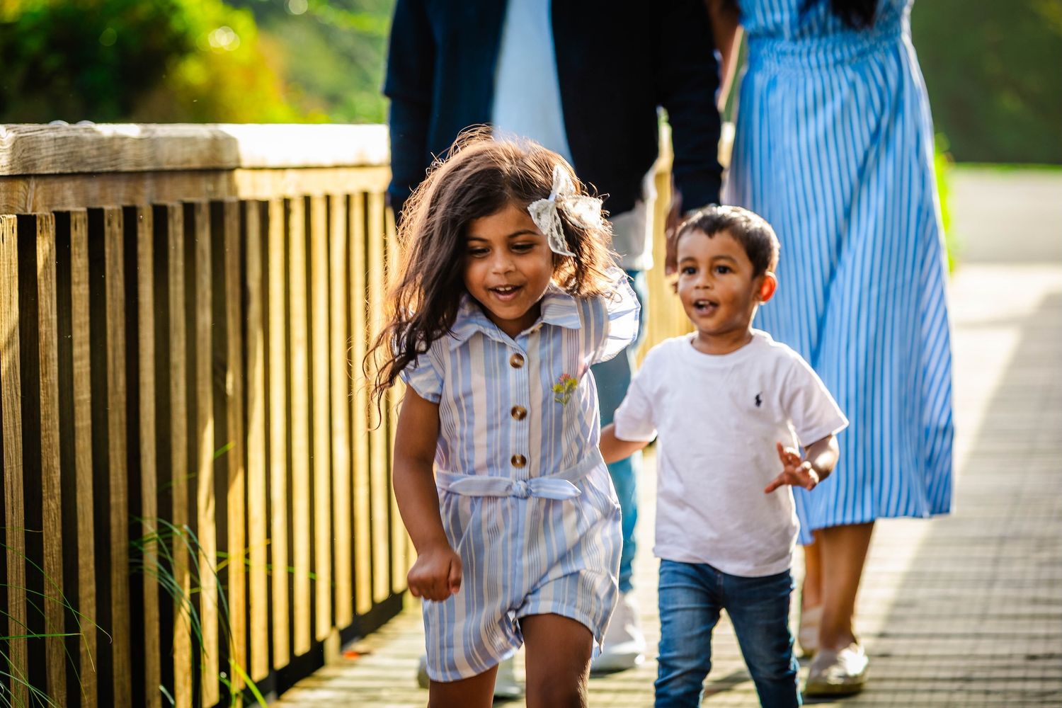 Two young children smile and walk on a wooden path outdoors, with adults in blue clothing visible behind them.