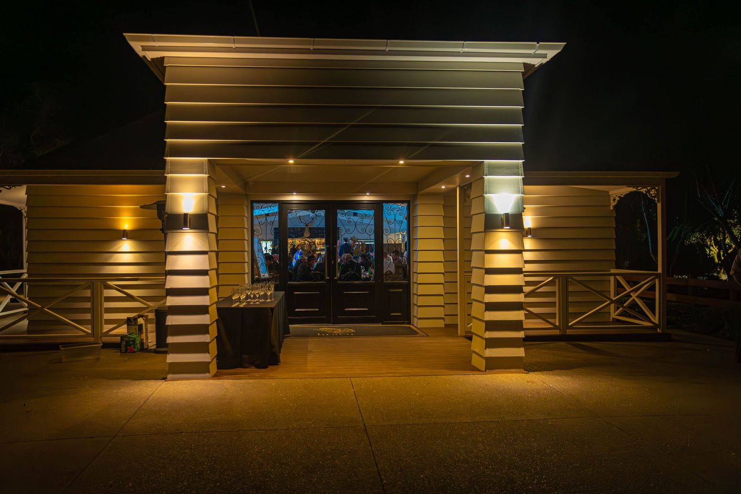 Illuminated restaurant entrance at night with warm wall lights flanking glass doors and white railings.
