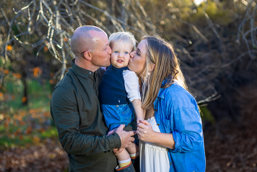 Parents kissing their baby boy on the cheeks outdoors in a sunlit park during a family portrait session.