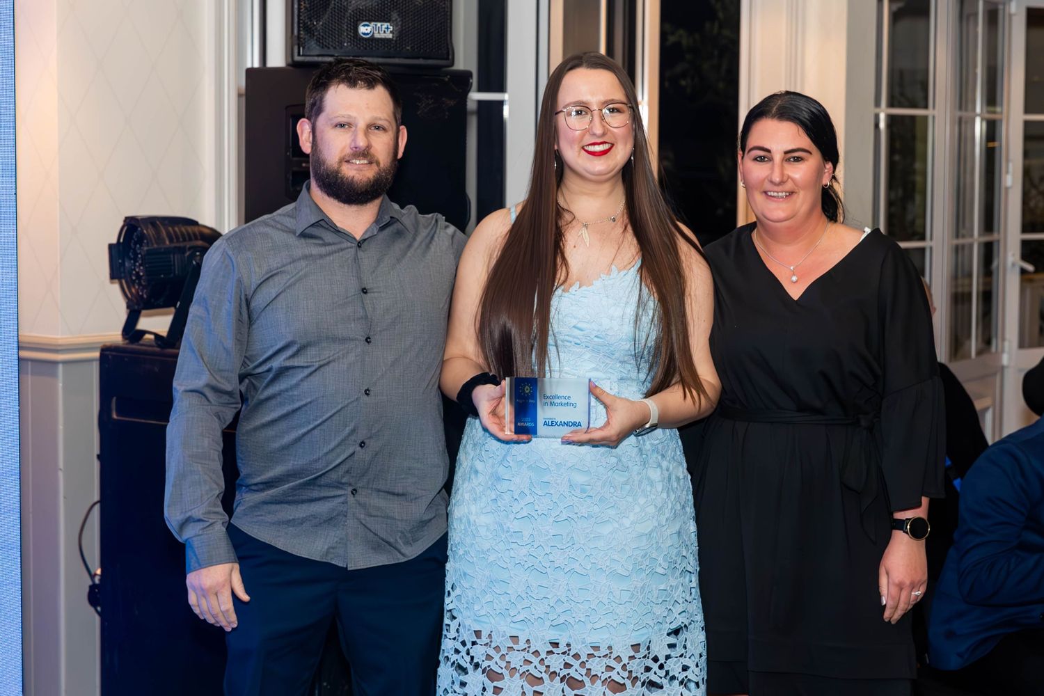 Three people posing together at an awards ceremony, with a woman in blue holding a trophy award plaque.
