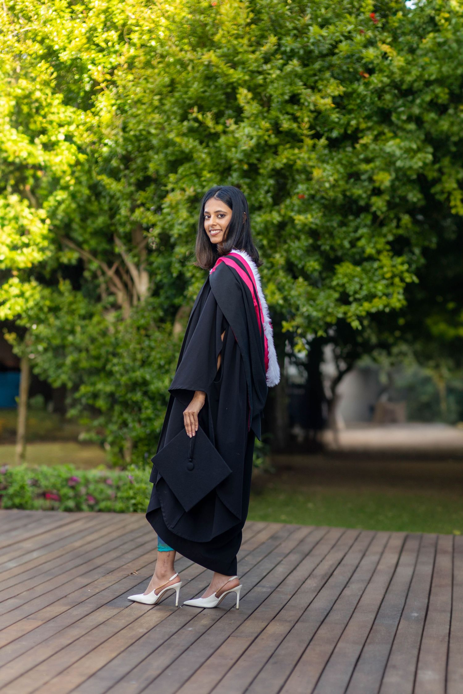 A young woman in black graduation gown and heels smiles over her shoulder outdoors on a wooden deck.