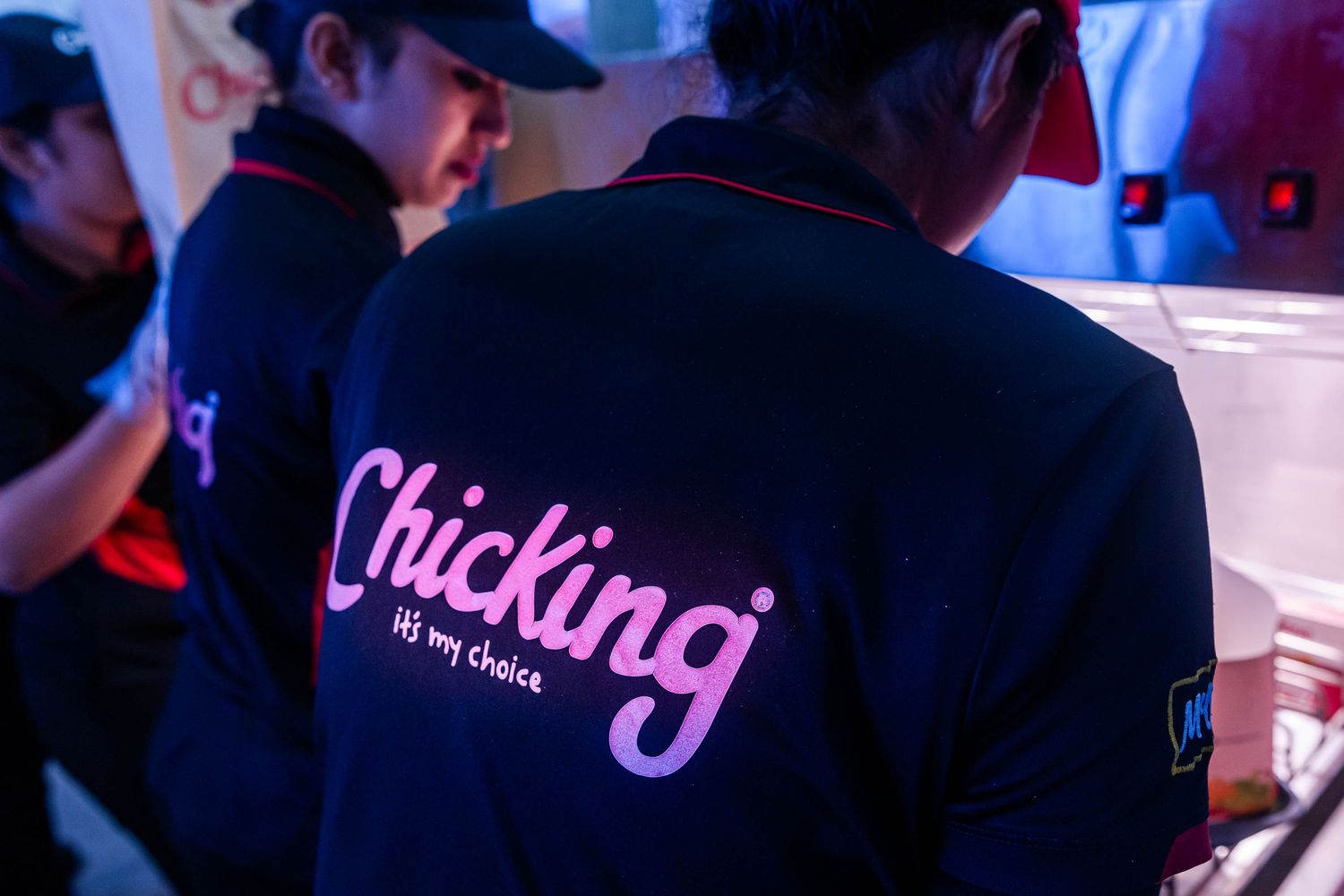 Chicking fast food restaurant staff member in branded navy uniform with pink logo, working at food preparation station.