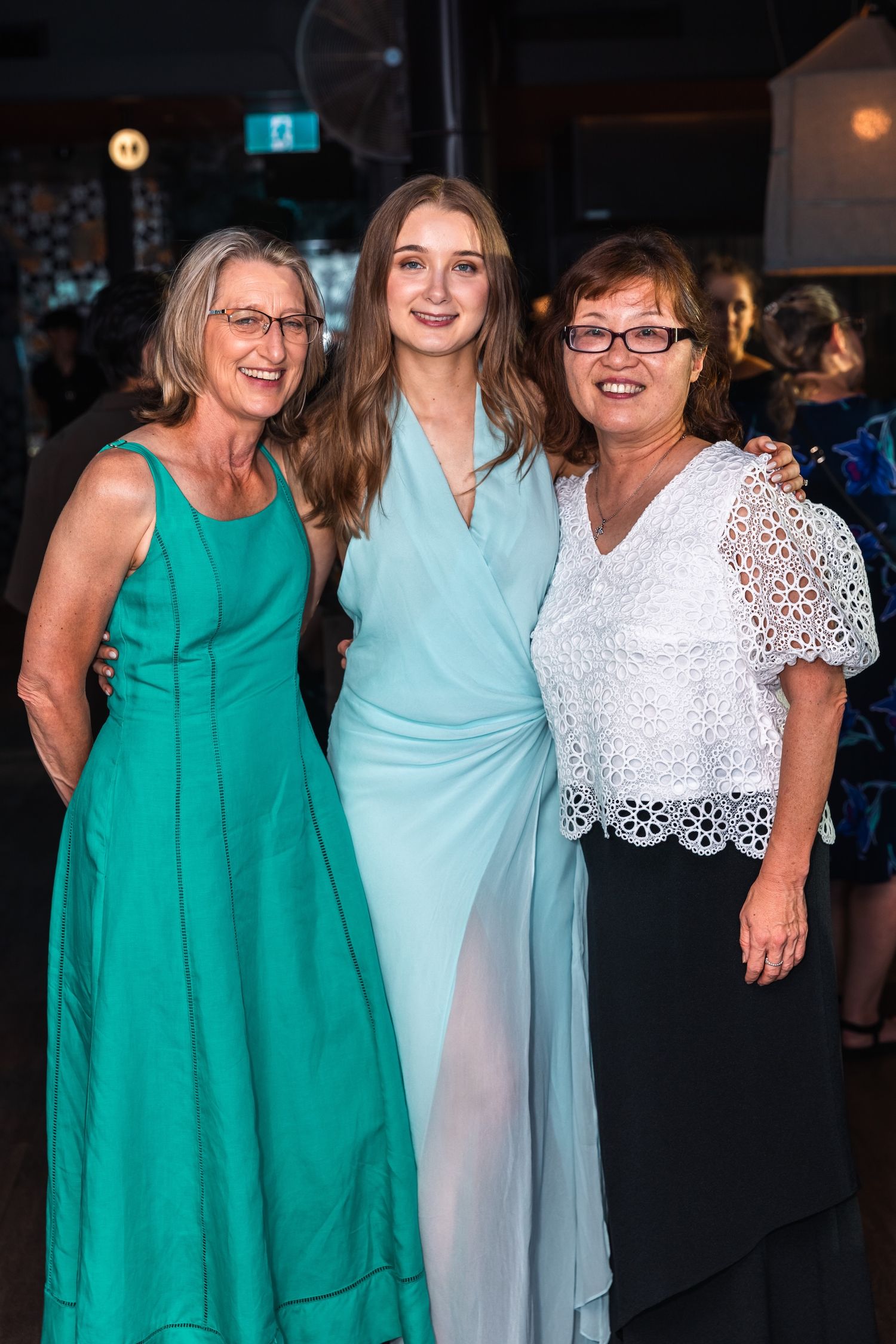 Three women in elegant formal gowns smile together at an event, wearing shades of teal, light blue, and white.