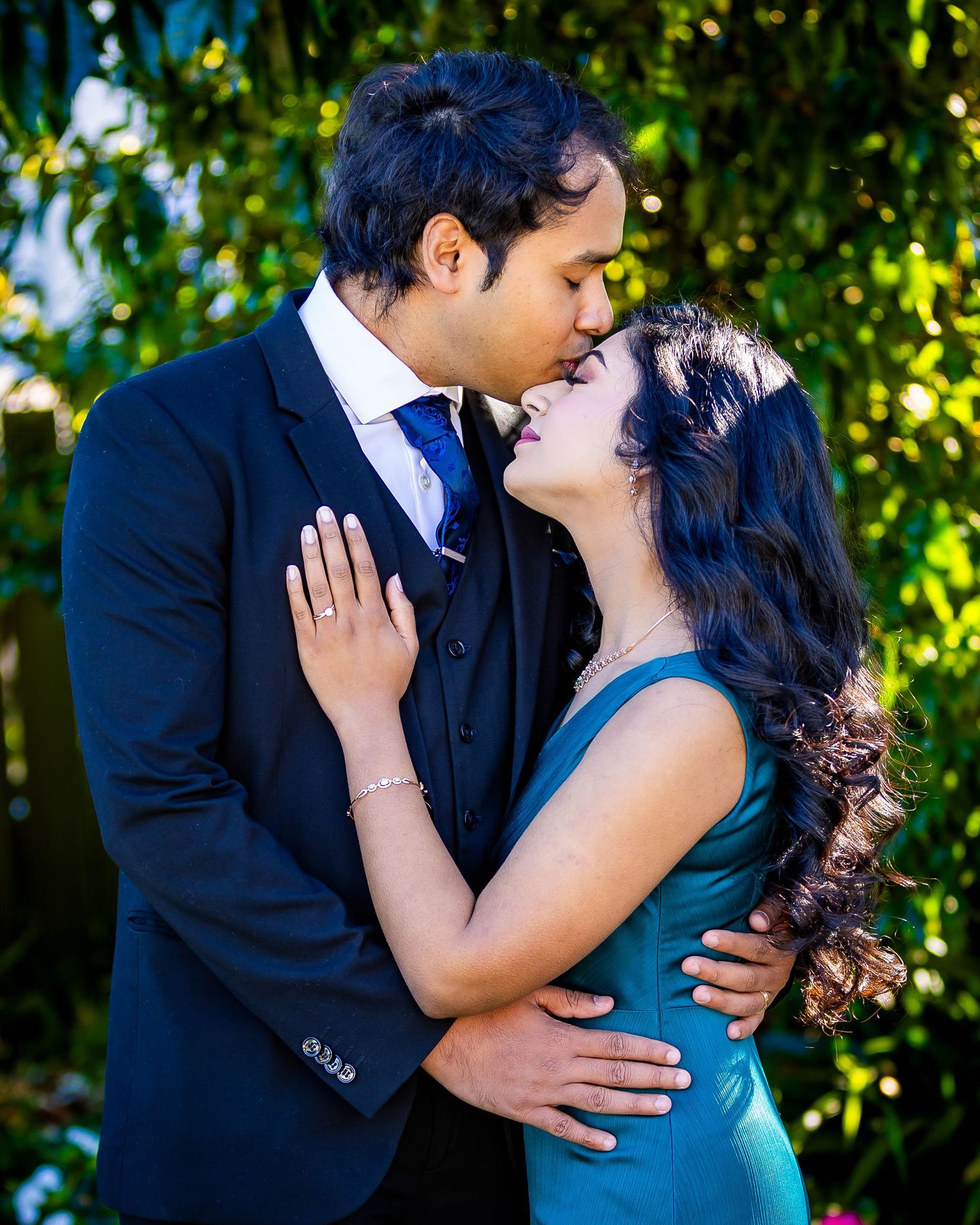 A couple shares a romantic kiss outdoors, man in navy suit, woman in teal dress, surrounded by lush green foliage.