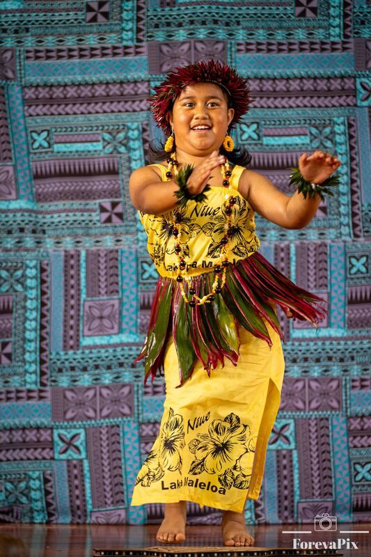 Young Polynesian girl in yellow traditional costume with leaf skirt and floral headpiece performing a cultural dance on stage.