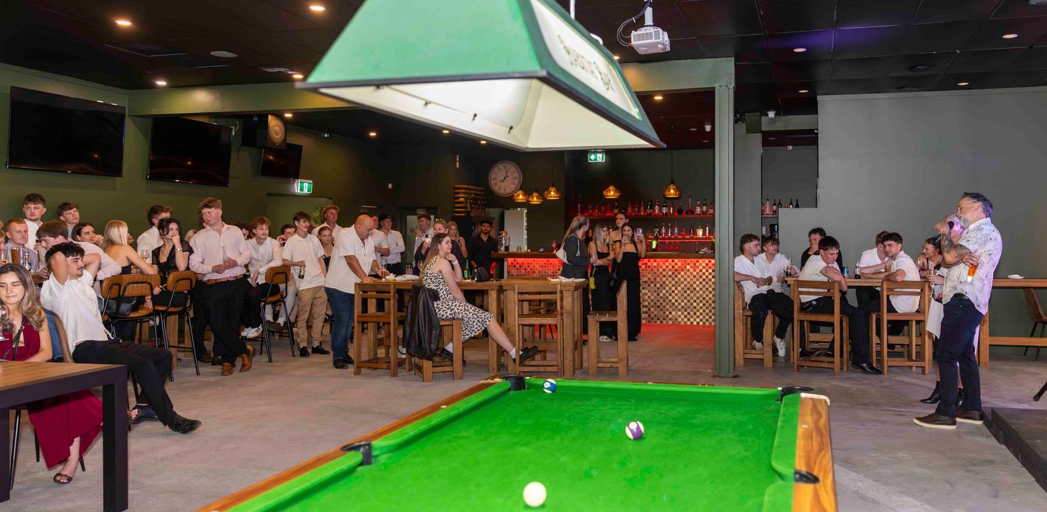Crowd watching a snooker game on a green billiard table in a bar with pendant lighting.