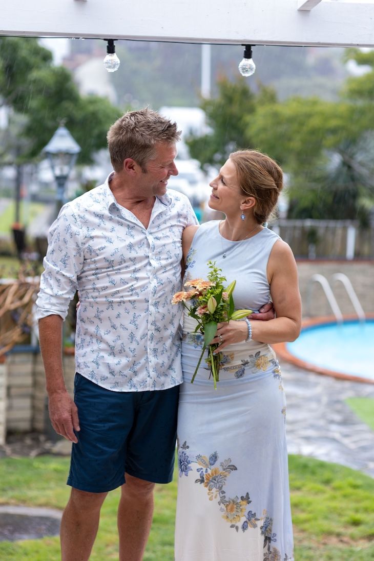 A couple shares a romantic moment outdoors, woman holding flowers in a light blue dress, man in a floral shirt near a pool.